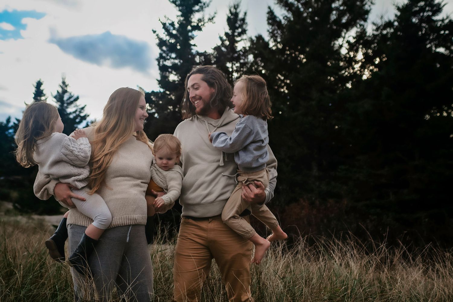 Berwick Happy family with two young children laughing together in a scenic meadow with pine trees at dusk.