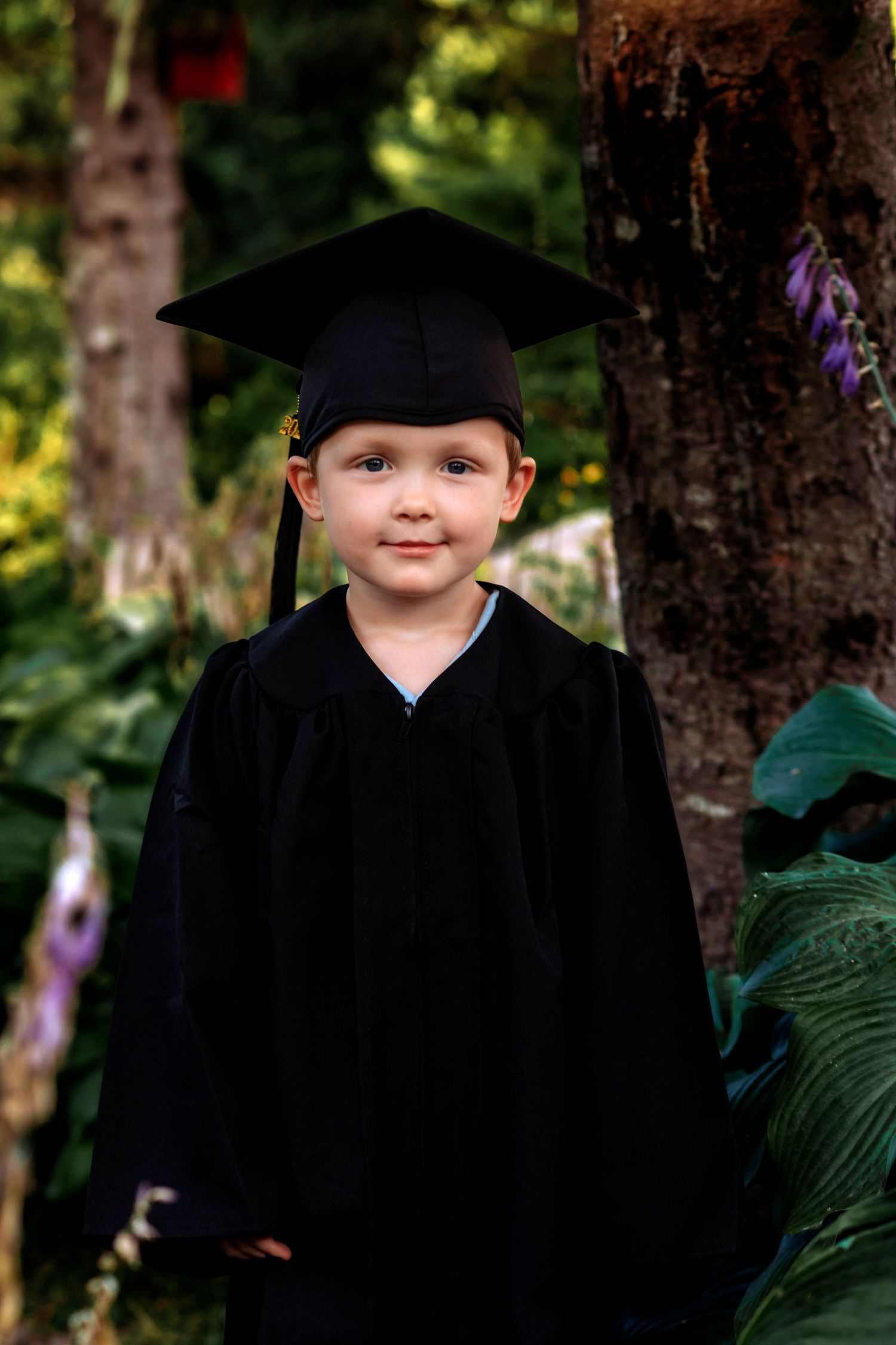 Young child wearing black graduation cap and gown smiling outdoors among green trees and plants.