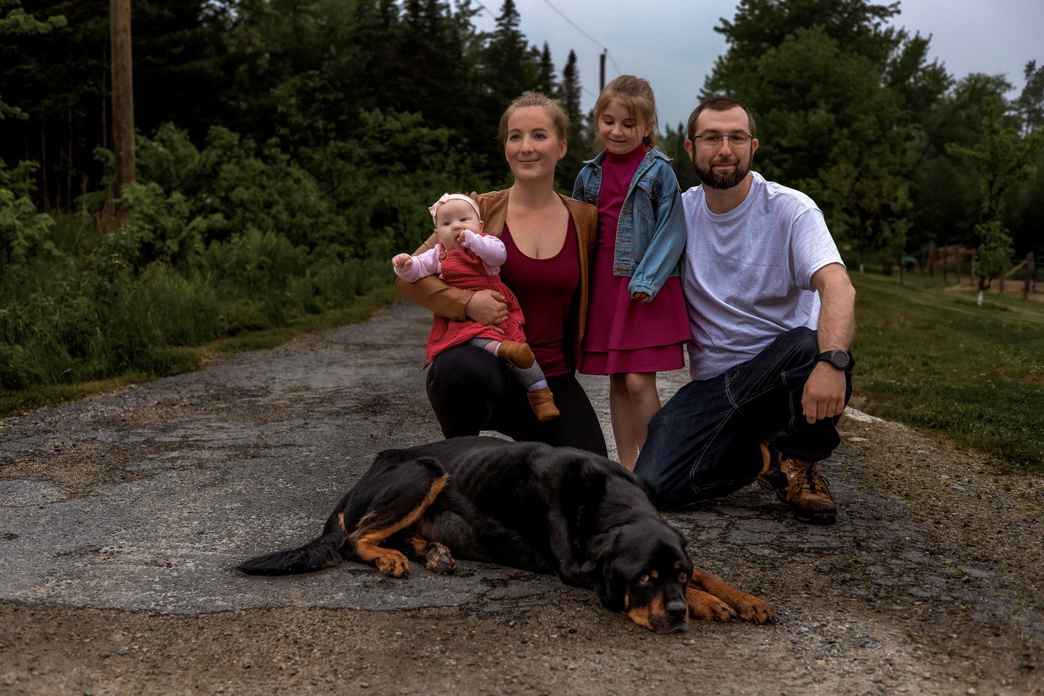 Family of three with baby poses beside a Rottweiler on a gravel path surrounded by green trees.