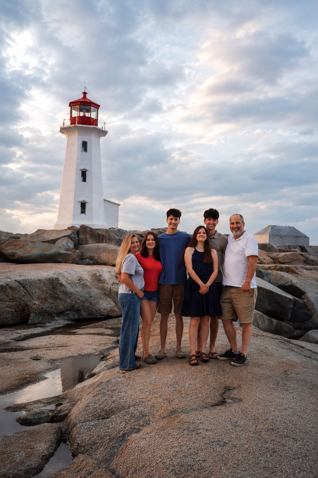 in Halifax area Family of five posing together on rocky shore with Peggy's Cove lighthouse and cloudy sky backdrop.