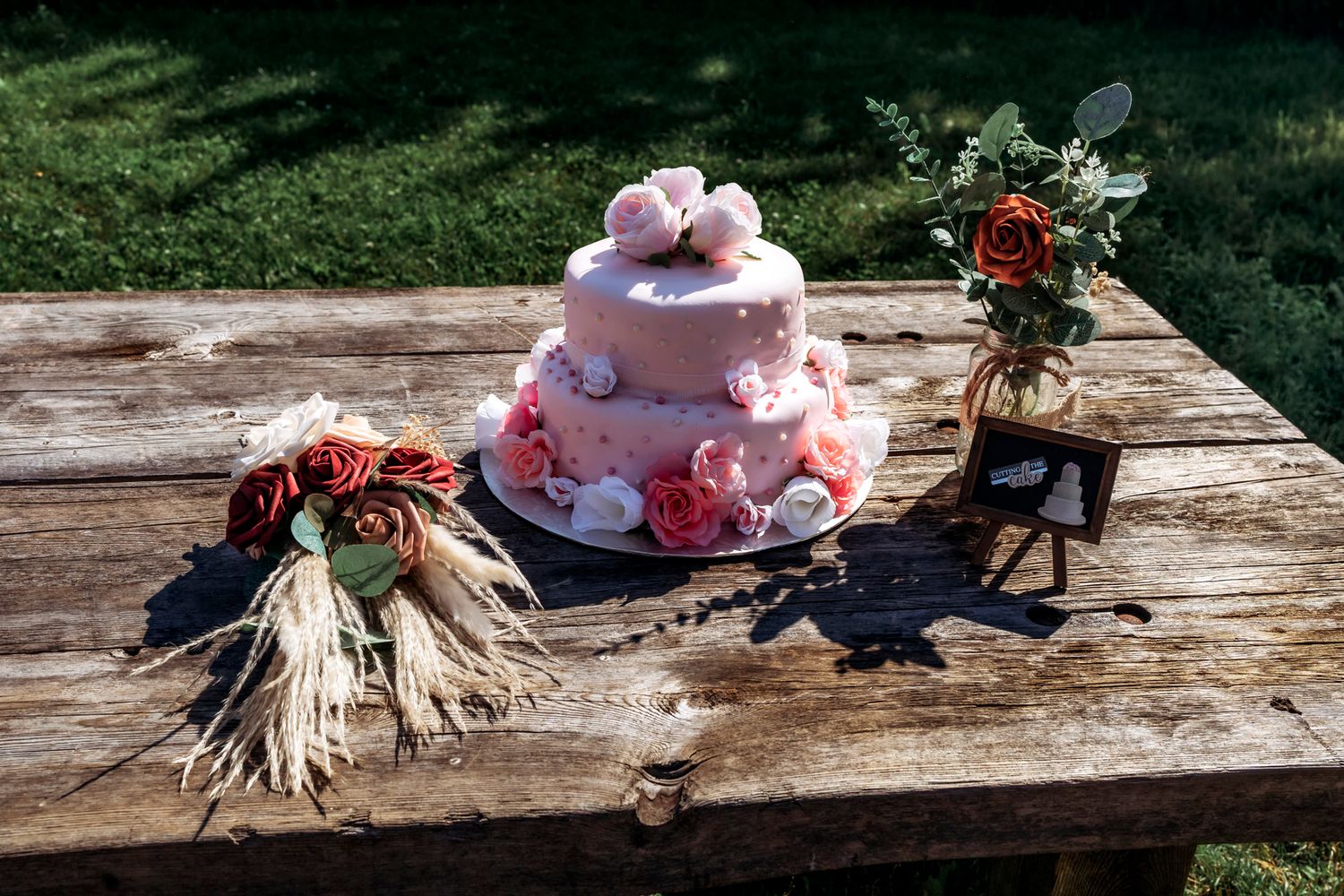 A two-tier pink wedding cake with floral decorations and dove toppers on a rustic wooden table outdoors.