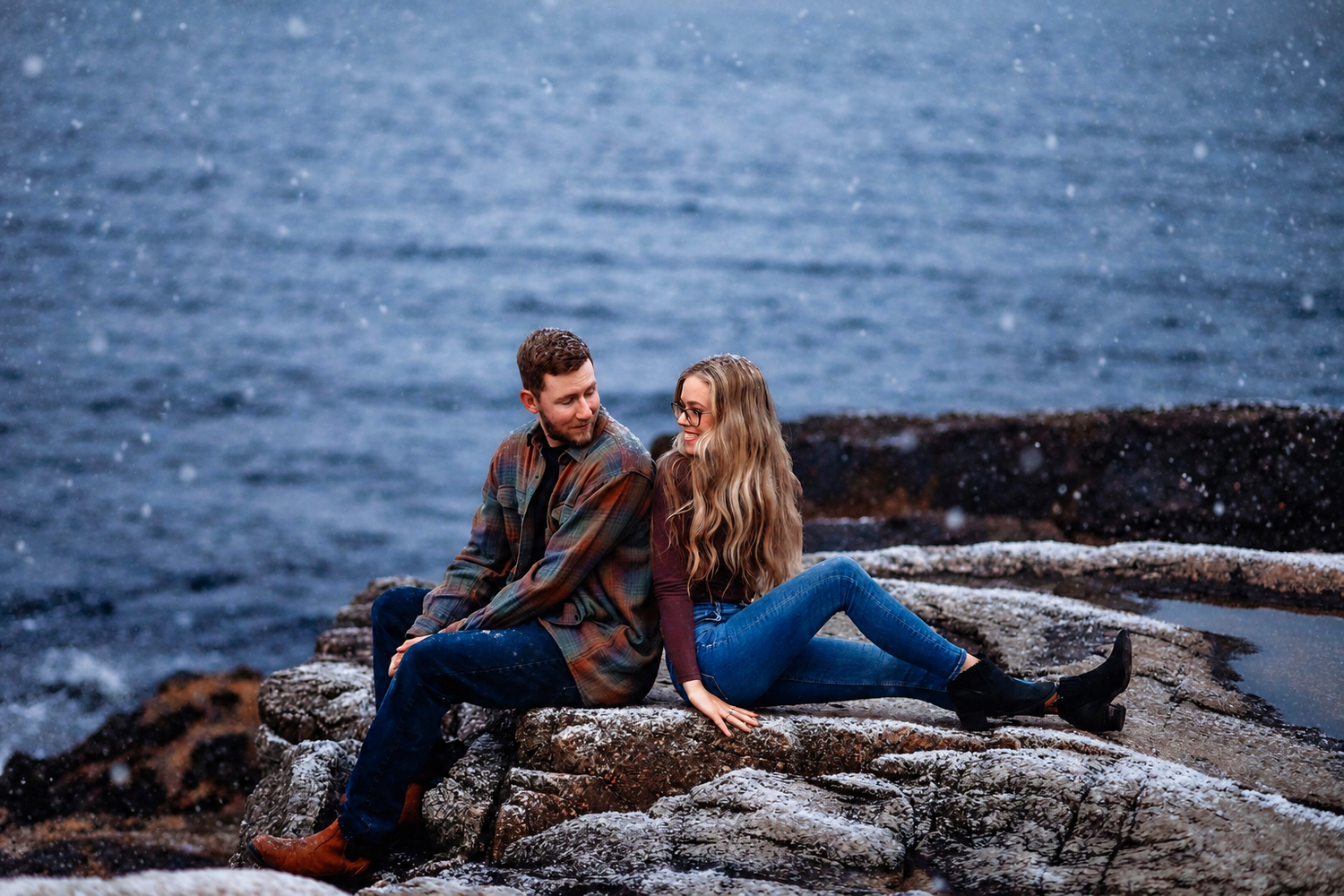 Couple in flannel jackets sitting together on rocky coastal cliffs overlooking the ocean while snow falls around them.