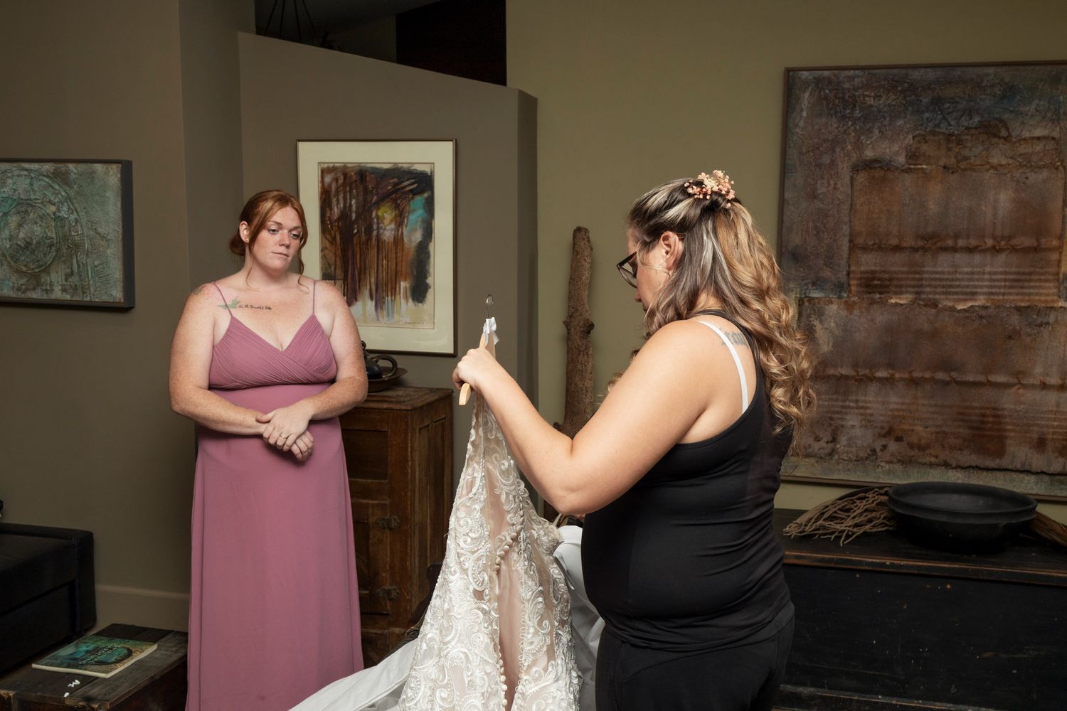 Bridesmaid in pink dress watches as woman in black holds up lace wedding gown in decorated room.