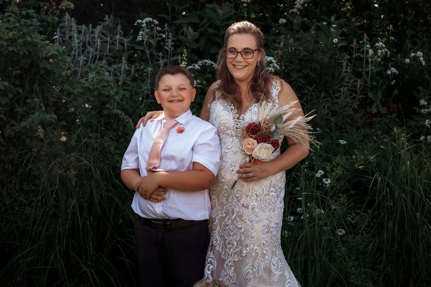 Bride in lace wedding gown holding bouquet poses with young boy in white shirt and tie outdoors.