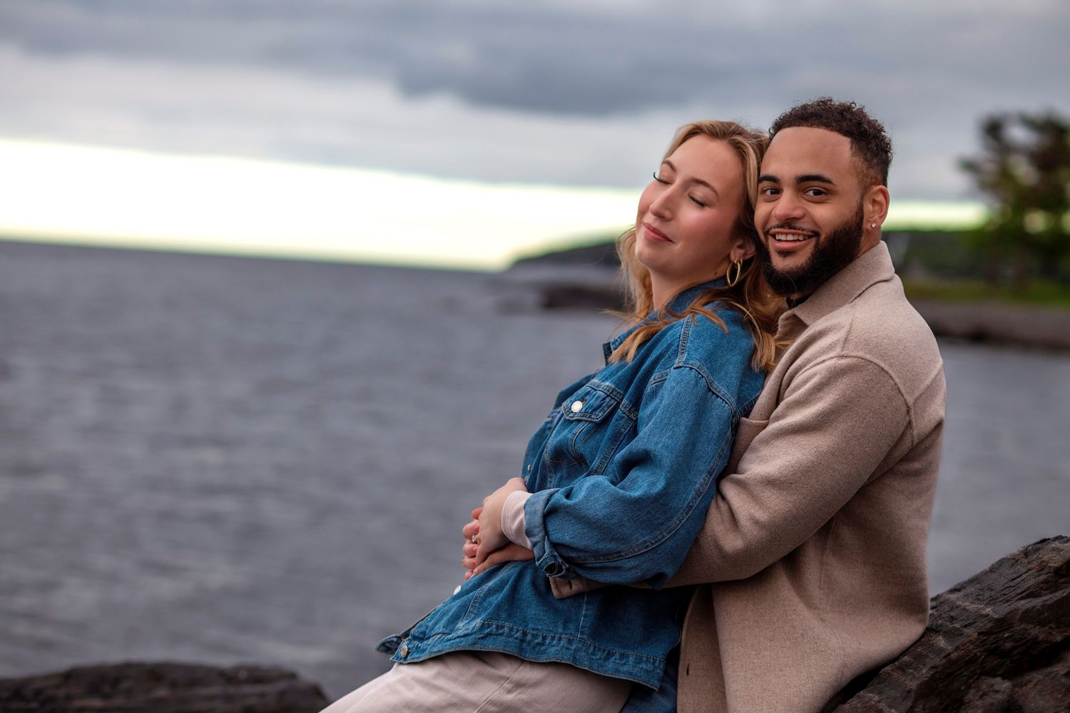 a Halifax Point pleasant park engagement session, a smiling interracial couple sits together on rocky shoreline