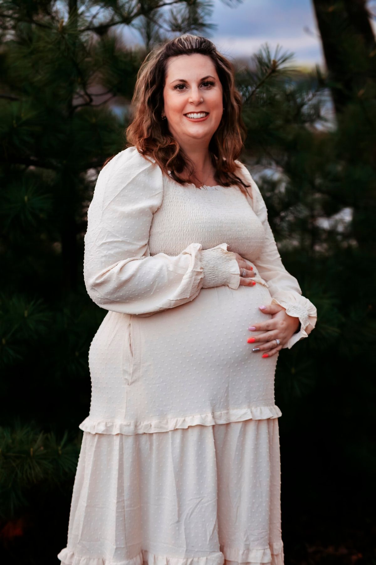 Smiling pregnant woman in white long-sleeve dress cradling baby bump outdoors among pine trees.