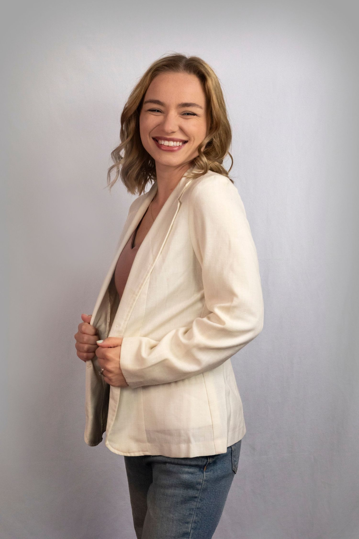 Smiling woman in cream blazer and jeans posing against gray background.