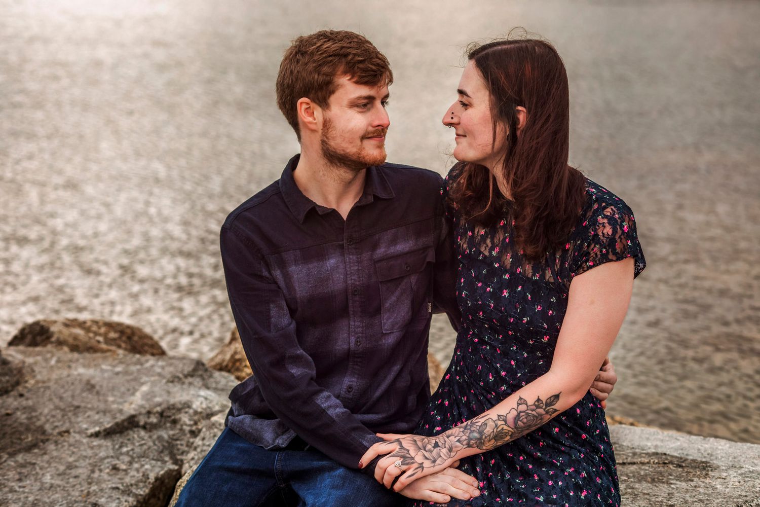 Couple sitting on rocks near water, smiling at each other, woman with tattoos wearing floral dress, man in plaid shirt.