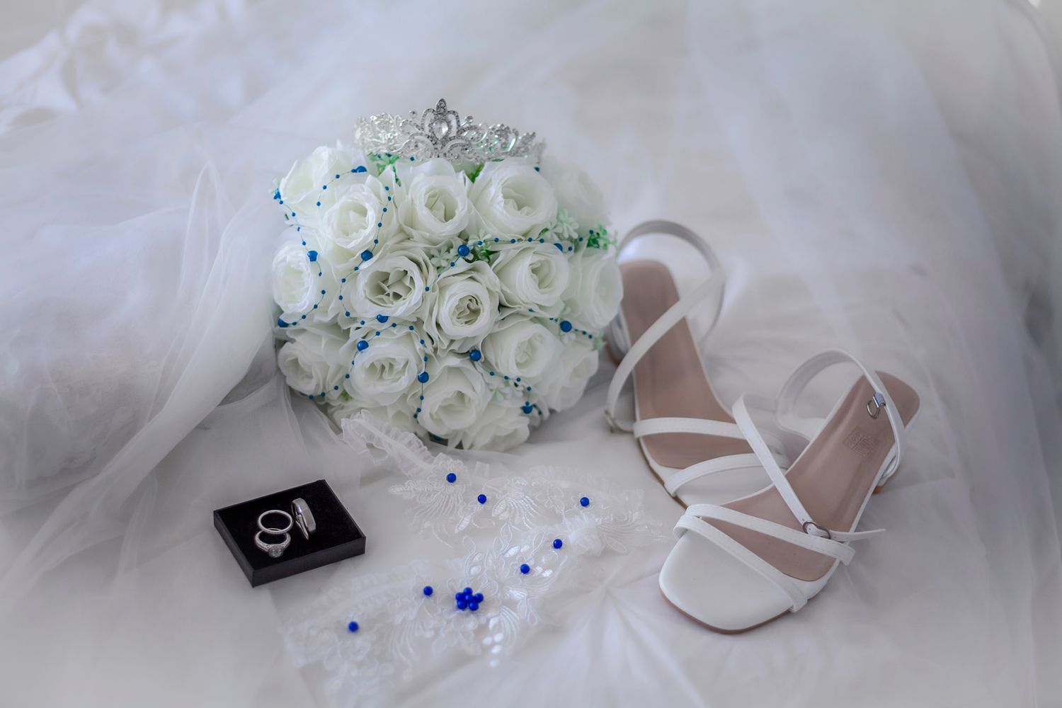 Elegant bridal accessories featuring white rose bouquet with tiara, strappy heels, rings, and blue accents on white fabric.