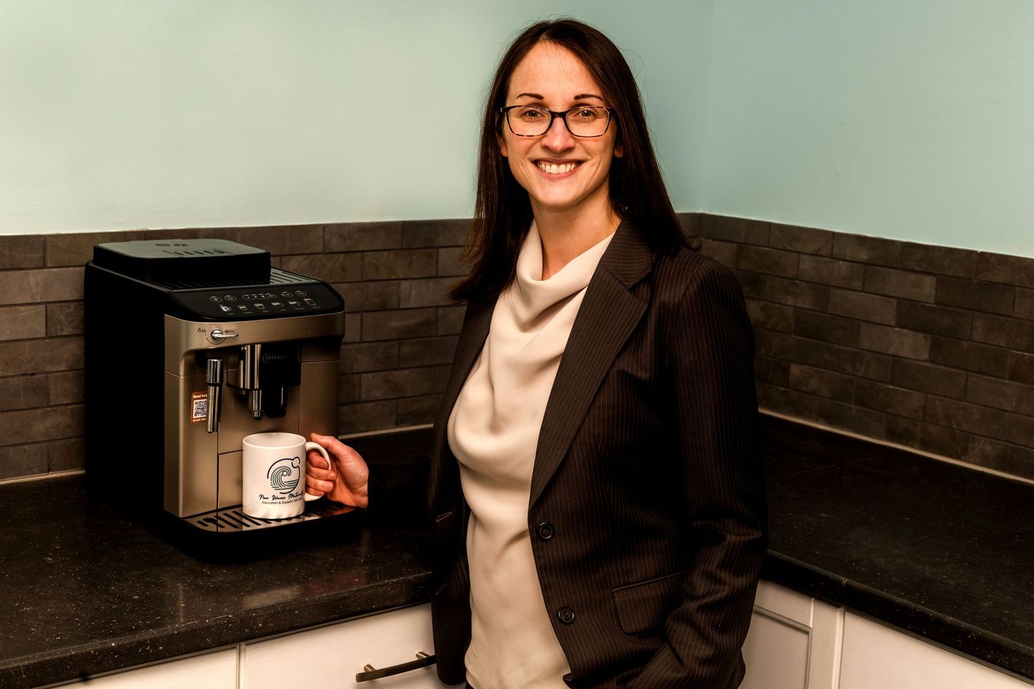 Professional woman in blazer smiling beside a modern espresso machine in a sleek office kitchen.
