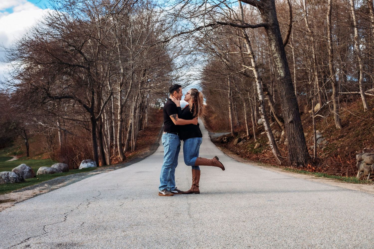 Couple sharing a romantic kiss on a quiet tree-lined road in autumn, woman lifting one leg in joy.