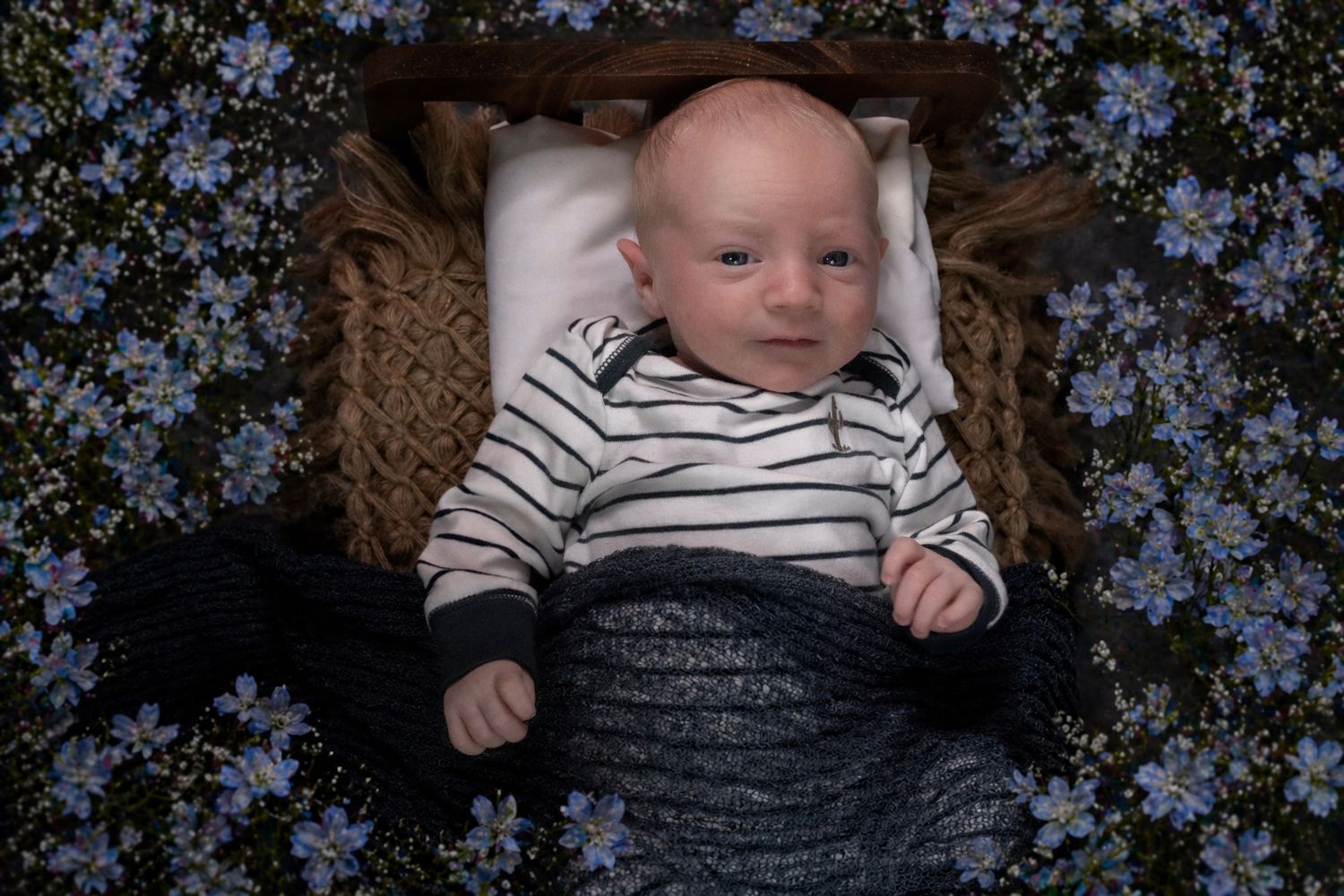Newborn baby in striped onesie lying in wicker basket surrounded by blue flowers.