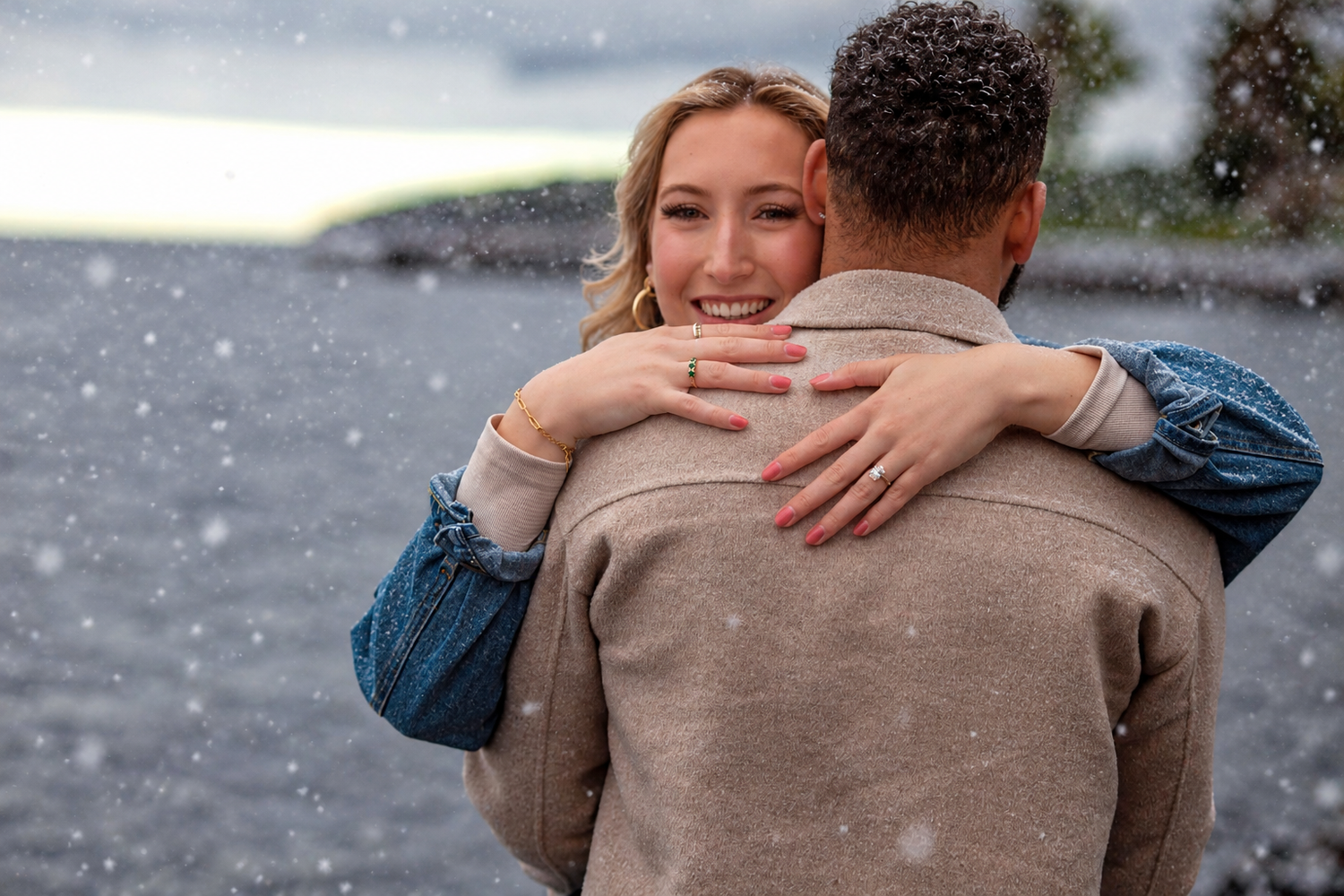 Smiling woman in a denim jacket embraces a man in a tan coat by a lake while snow falls around them in a romantic winter engagement moment.