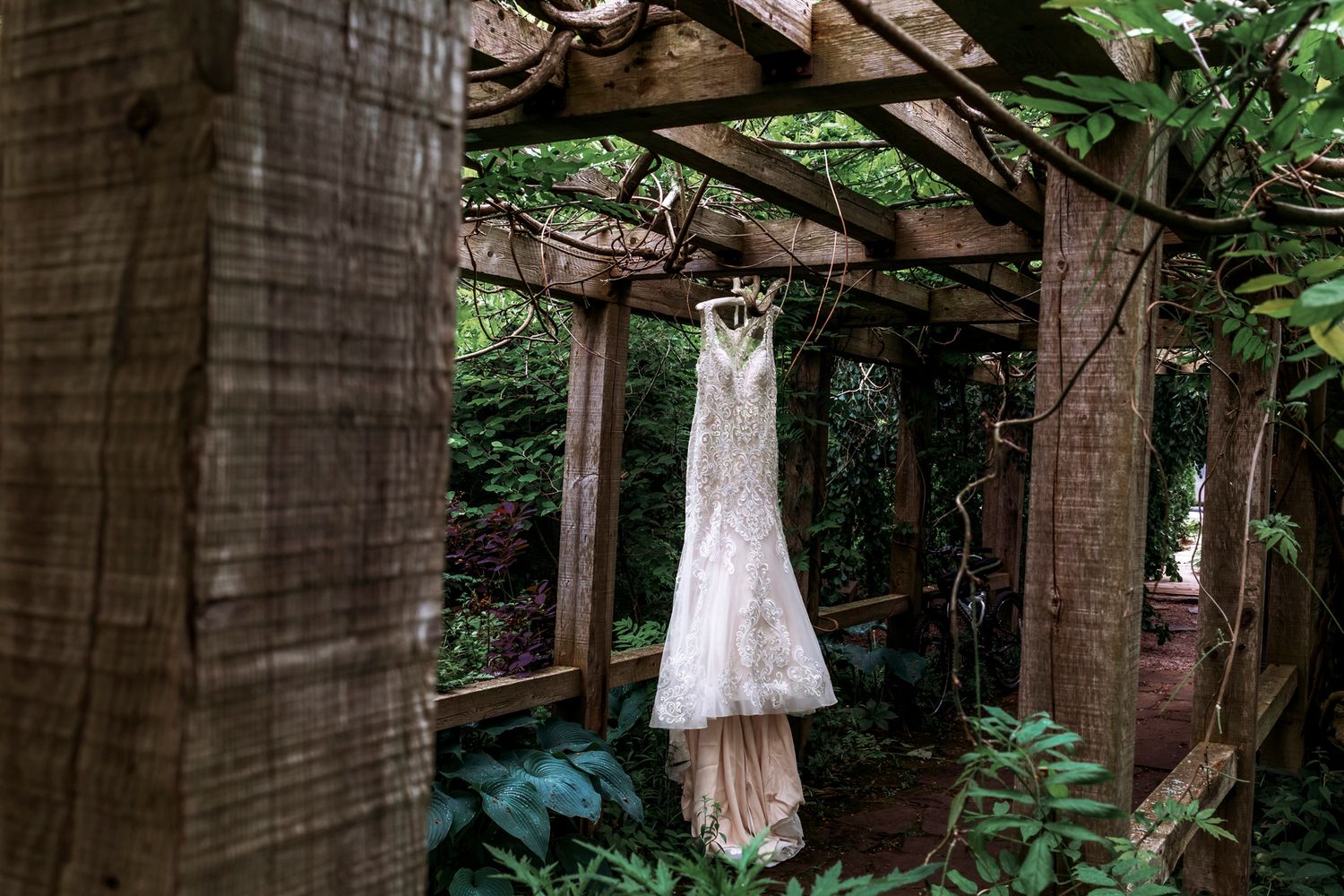 A lace wedding dress hanging in a rustic wooden garden pergola surrounded by lush green foliage.