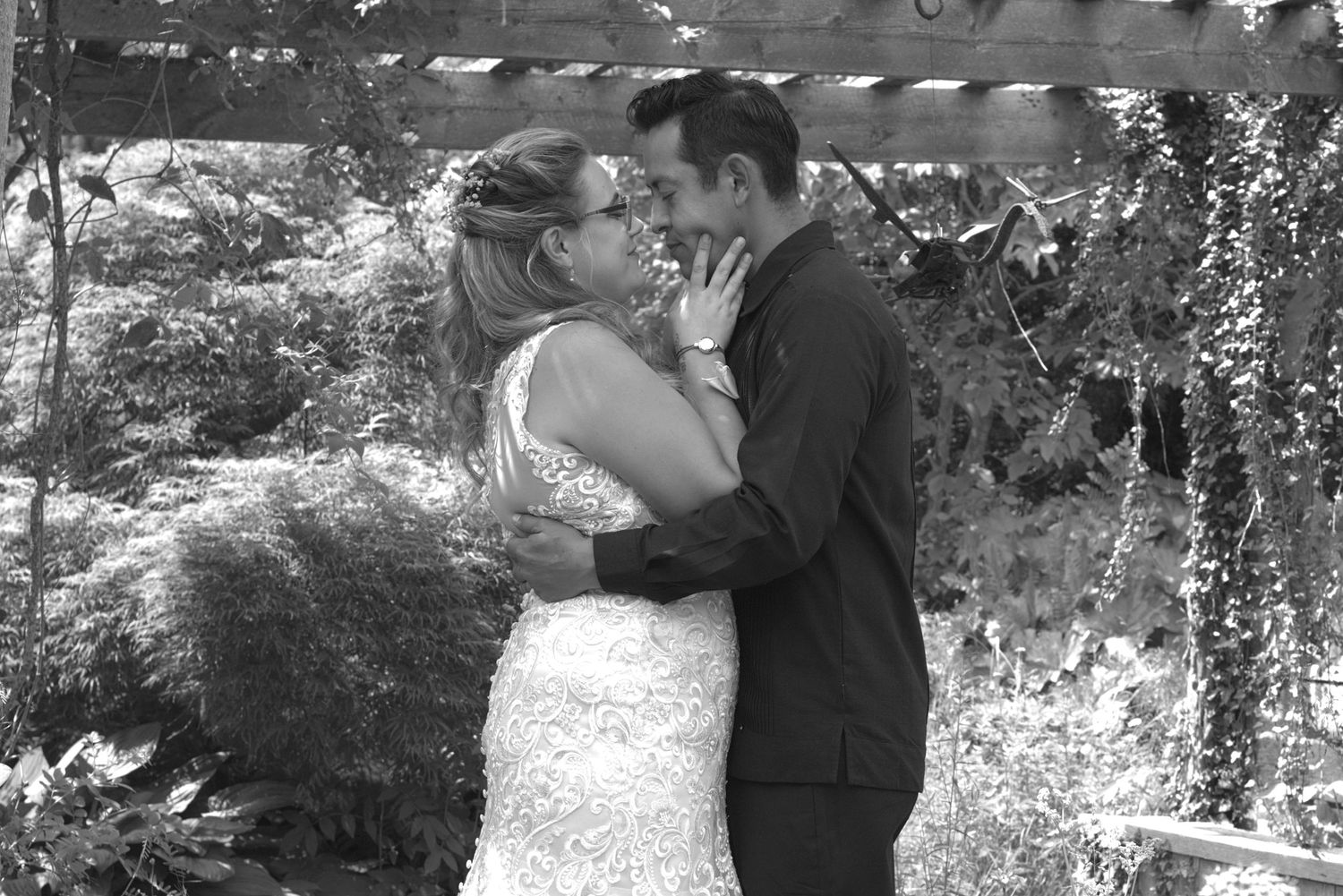 Black and white photo of a bride and groom sharing a romantic moment under a garden arbor on their wedding day.