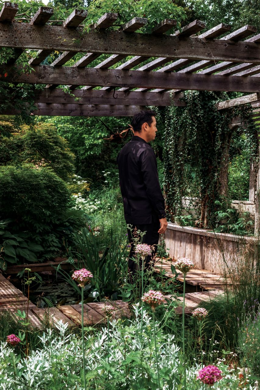 Man in black outfit standing in a lush garden under a wooden pergola surrounded by pink flowers and greenery.