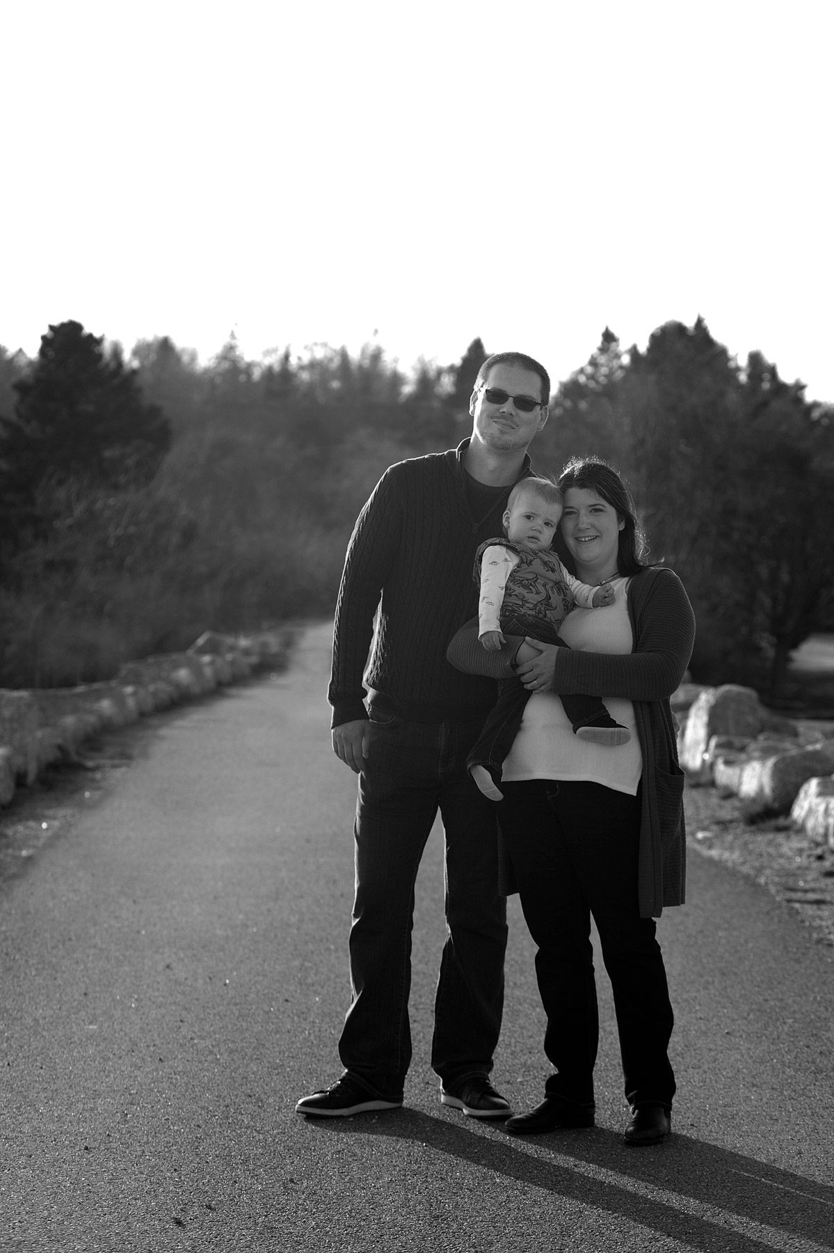 Annapolis Valley Family Photographer, Black and white photo of a couple standing on a road holding their baby, trees in the background.
