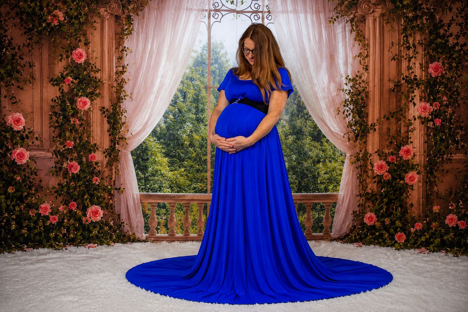 Pregnant woman in royal blue maternity gown posing before floral backdrop with pink roses and sheer curtains.