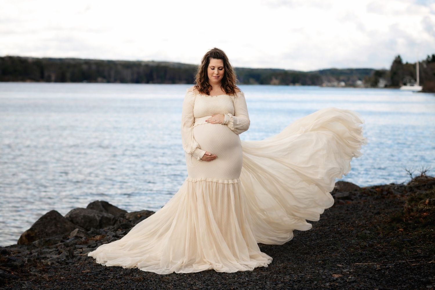 Halifax photographer, Pregnant woman in flowing cream gown poses by lakeside on rocks, dress billowing in wind during maternity photoshoot.