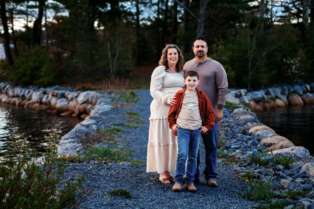 Family of three posing on a rocky waterfront path at dusk, mother in cream dress, father in gray, son in striped shirt.