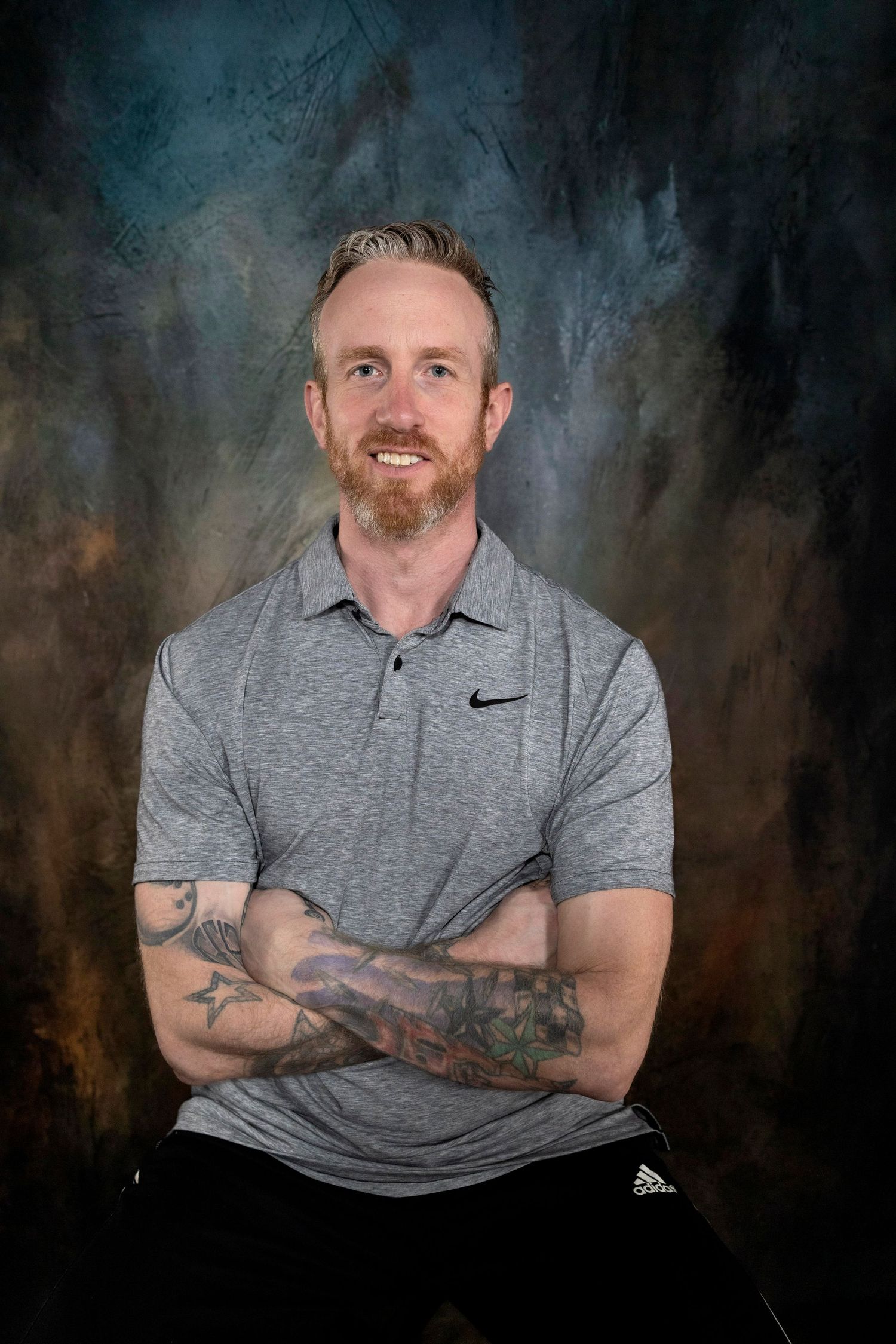 Smiling man with red beard, tattoos, and crossed arms wearing a gray Nike polo shirt against a textured backdrop.