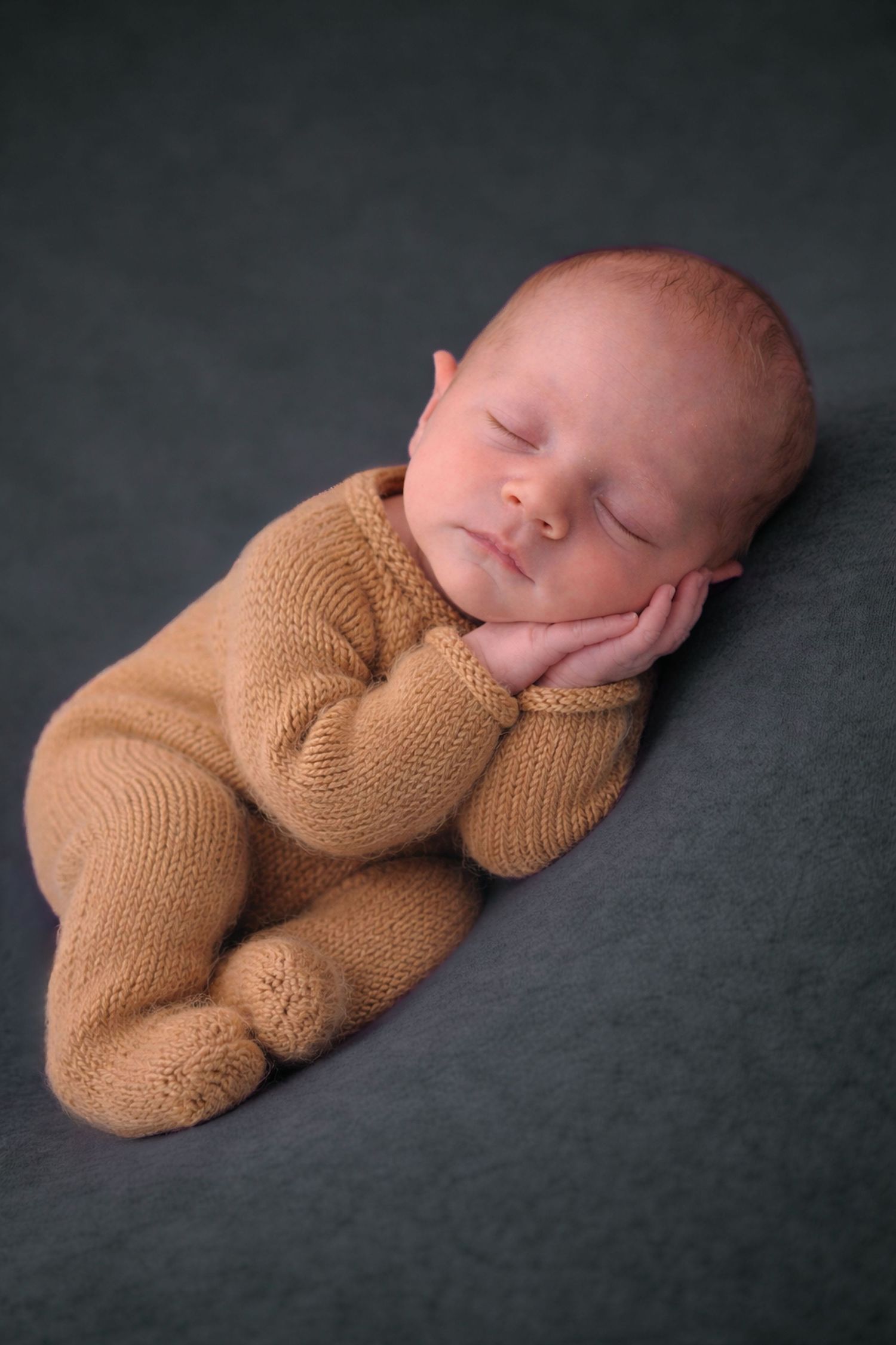 Peaceful newborn baby sleeping in a tan knit romper on a gray background.