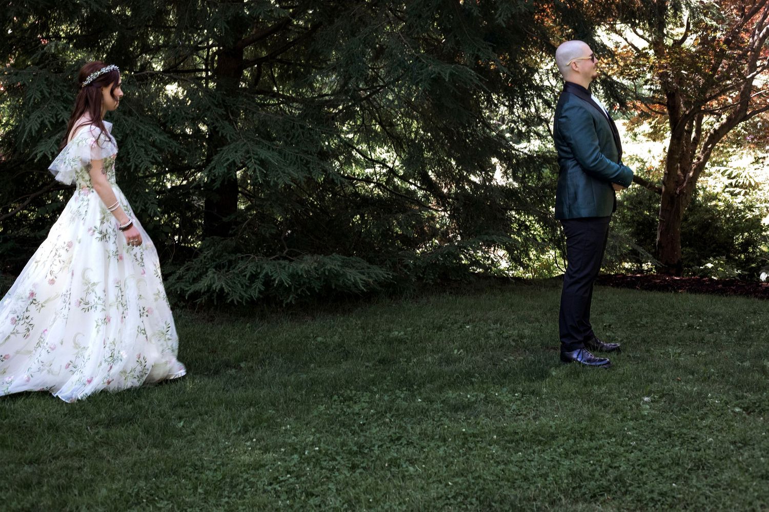 Bride in floral gown faces groom in black suit on green lawn surrounded by tall evergreen trees.