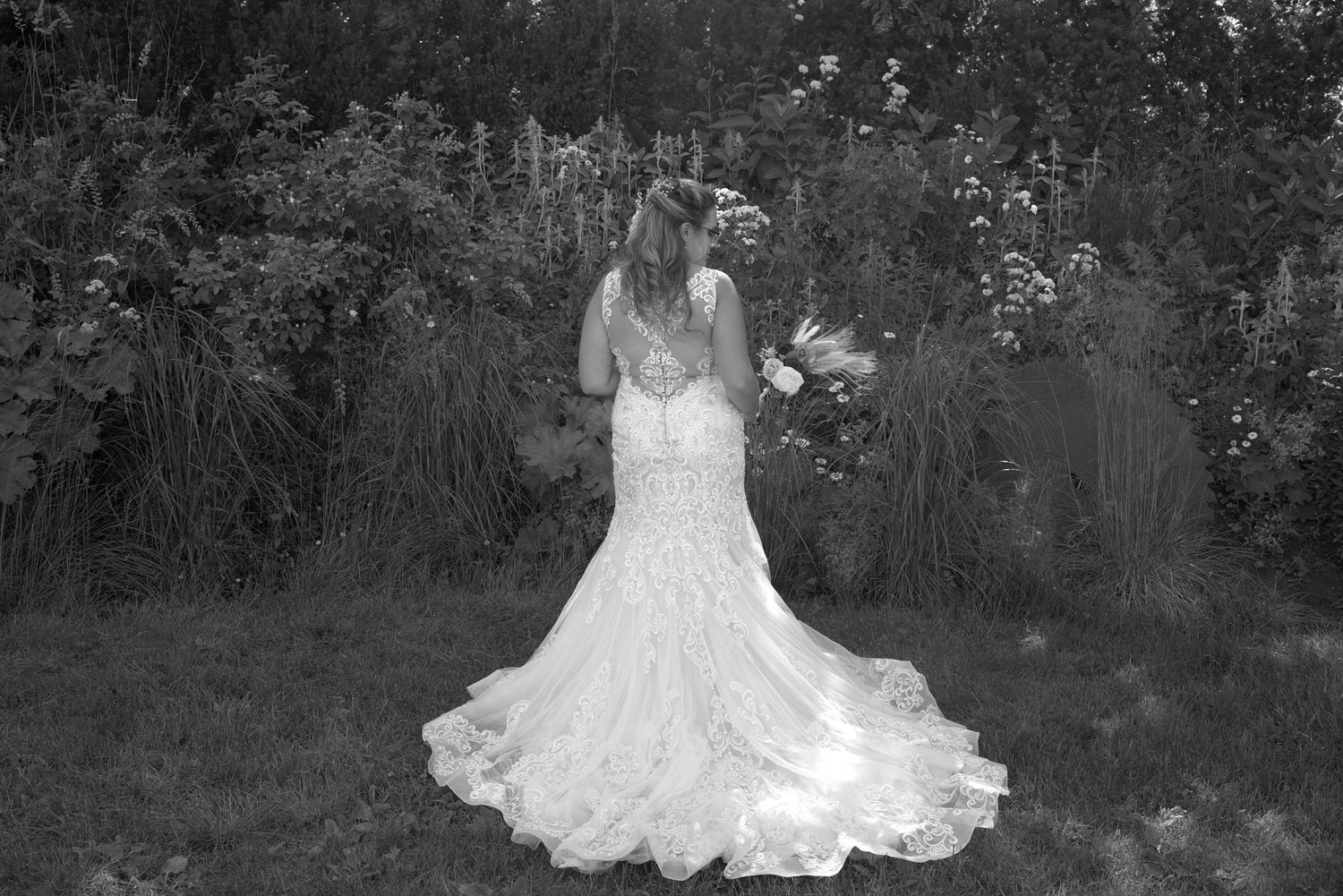 Black and white photo of a bride in a lace wedding gown with floral crown holding bouquet in a garden.