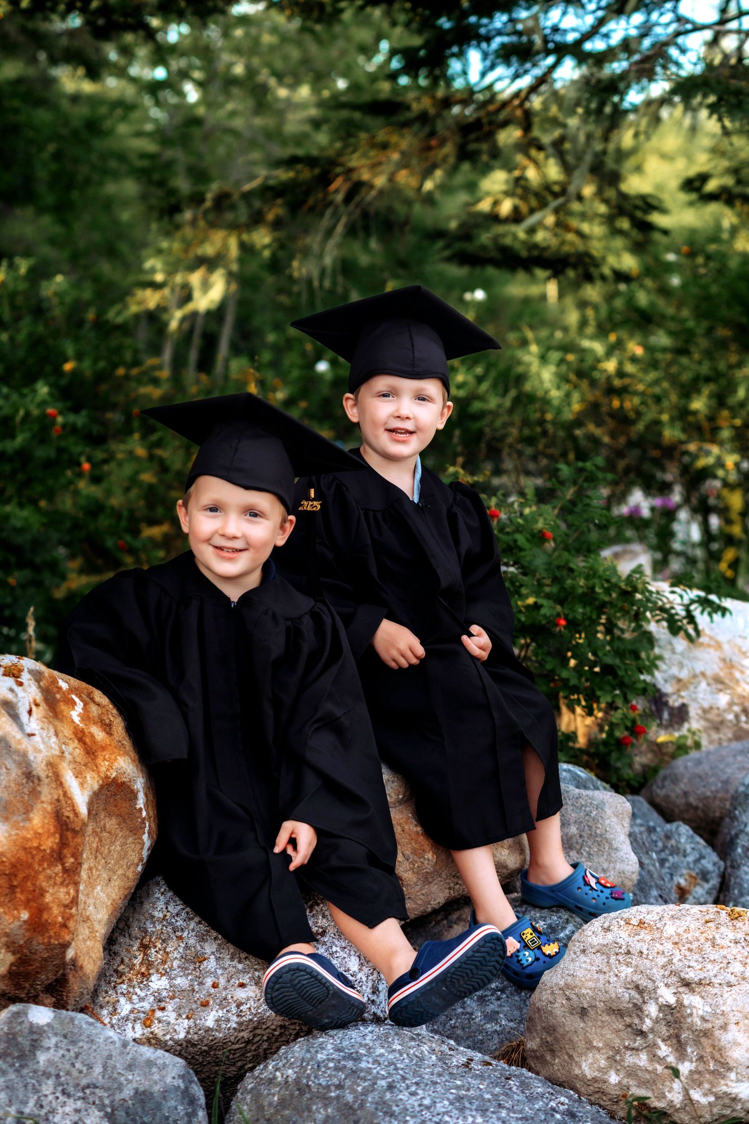 Two young children in black graduation caps and gowns sitting on rocks outdoors, smiling for a graduation photo.
