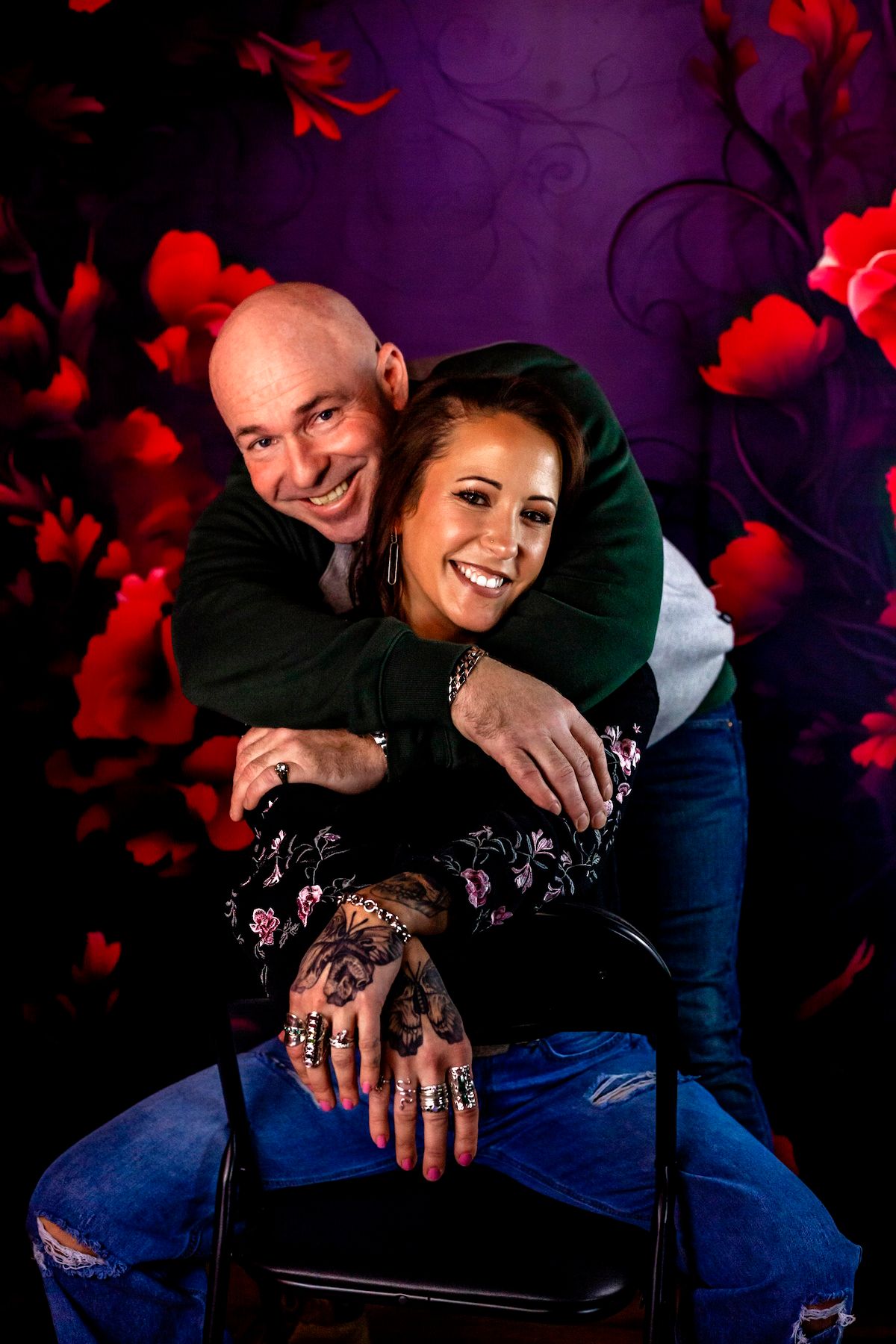 A bald man hugs a smiling tattooed woman with rings, posed before a red floral purple studio backdrop.