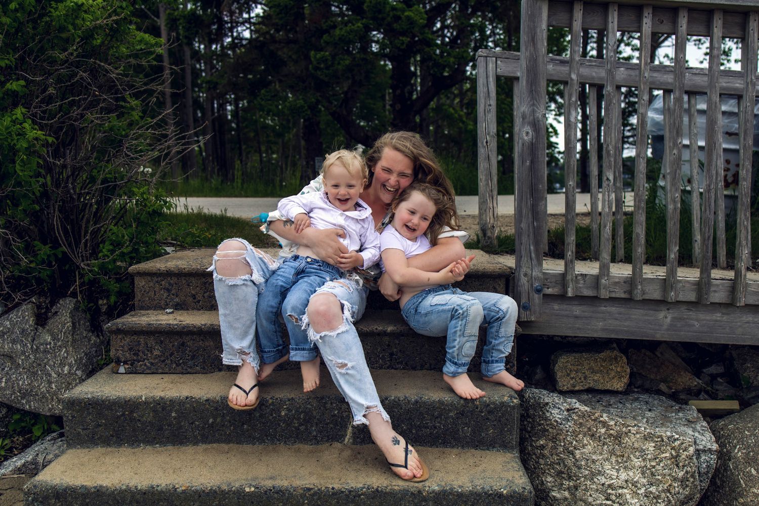 in Windsor a Woman sitting on stone steps with two young children in matching ripped jeans, smiling outdoors near wooden deck.