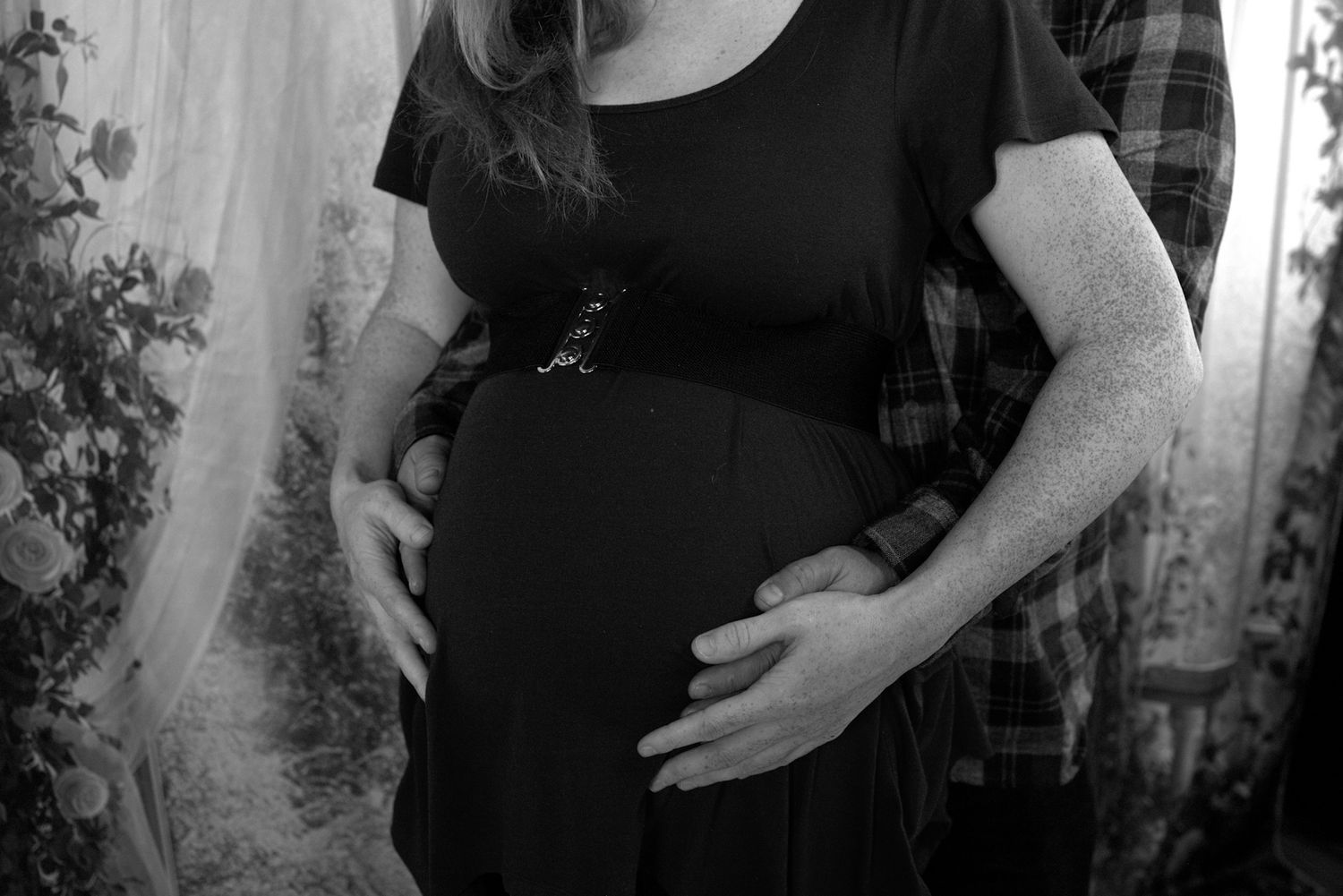 Black and white photo of a pregnant woman in a black dress with her partner's hands on her baby bump.