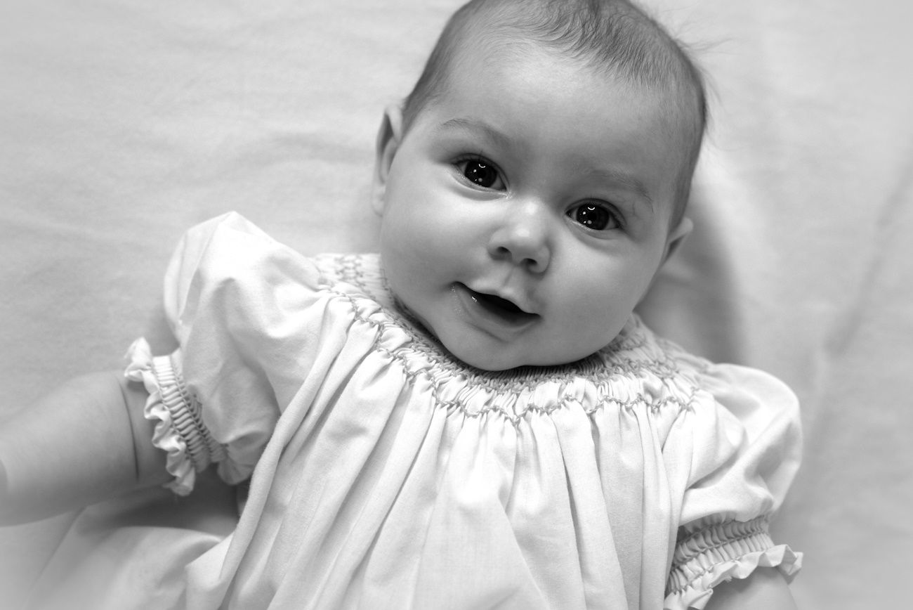Halifax newborn photographer Black and white portrait of a smiling baby in a white smocked dress lying on a soft white blanket.