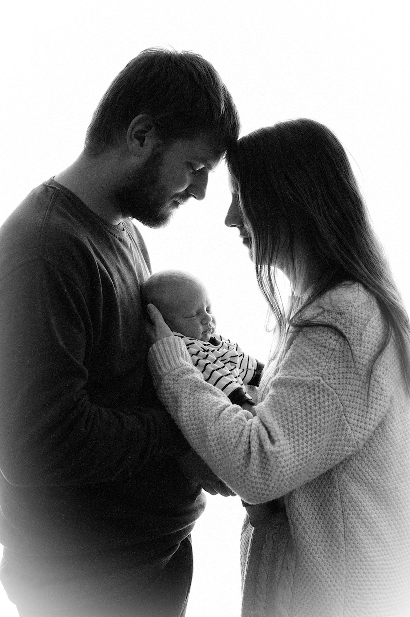 Black and white photo of parents tenderly holding their newborn baby, foreheads touching in an intimate family portrait.