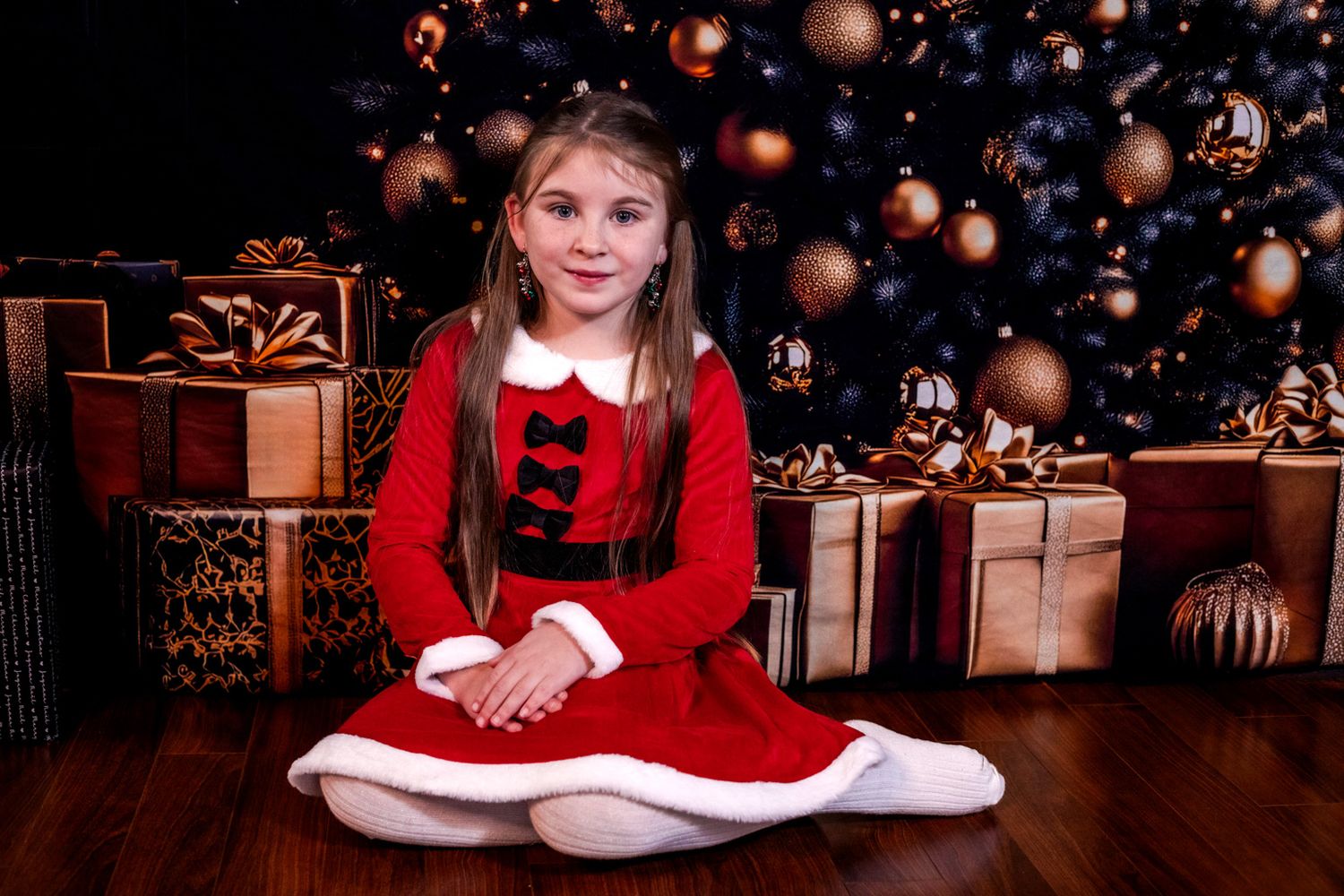 Smiling girl in a red Santa dress sits on a wooden floor beside wrapped gifts and a Christmas tree with gold ornaments.