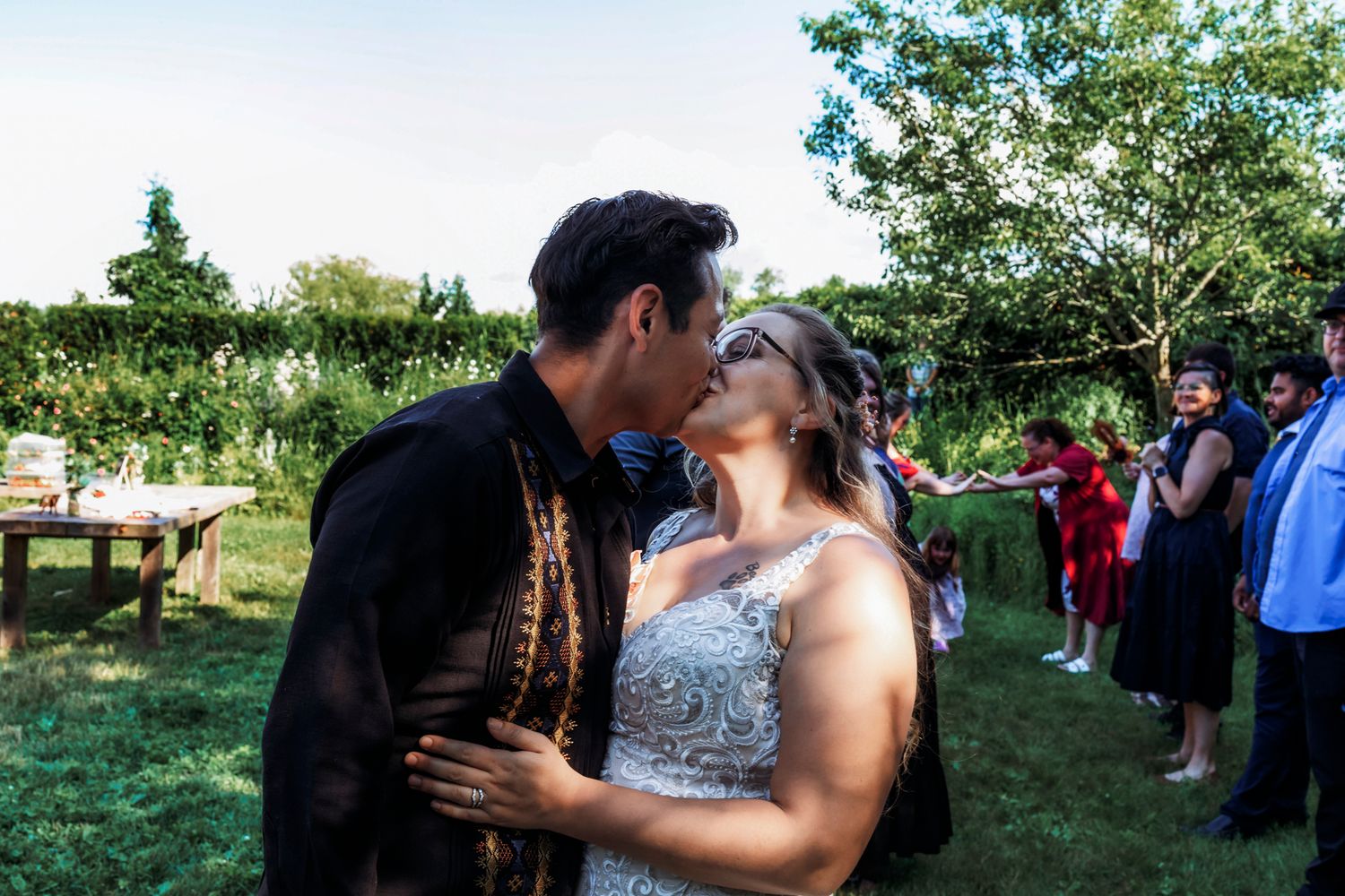 A couple shares a romantic kiss during an outdoor wedding ceremony, surrounded by guests in a lush green garden.