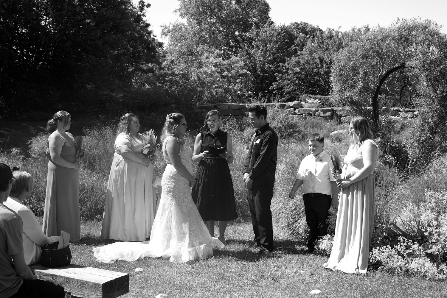 Black and white outdoor wedding ceremony with bride, groom, officiant, and wedding party standing on grass surrounded by trees.