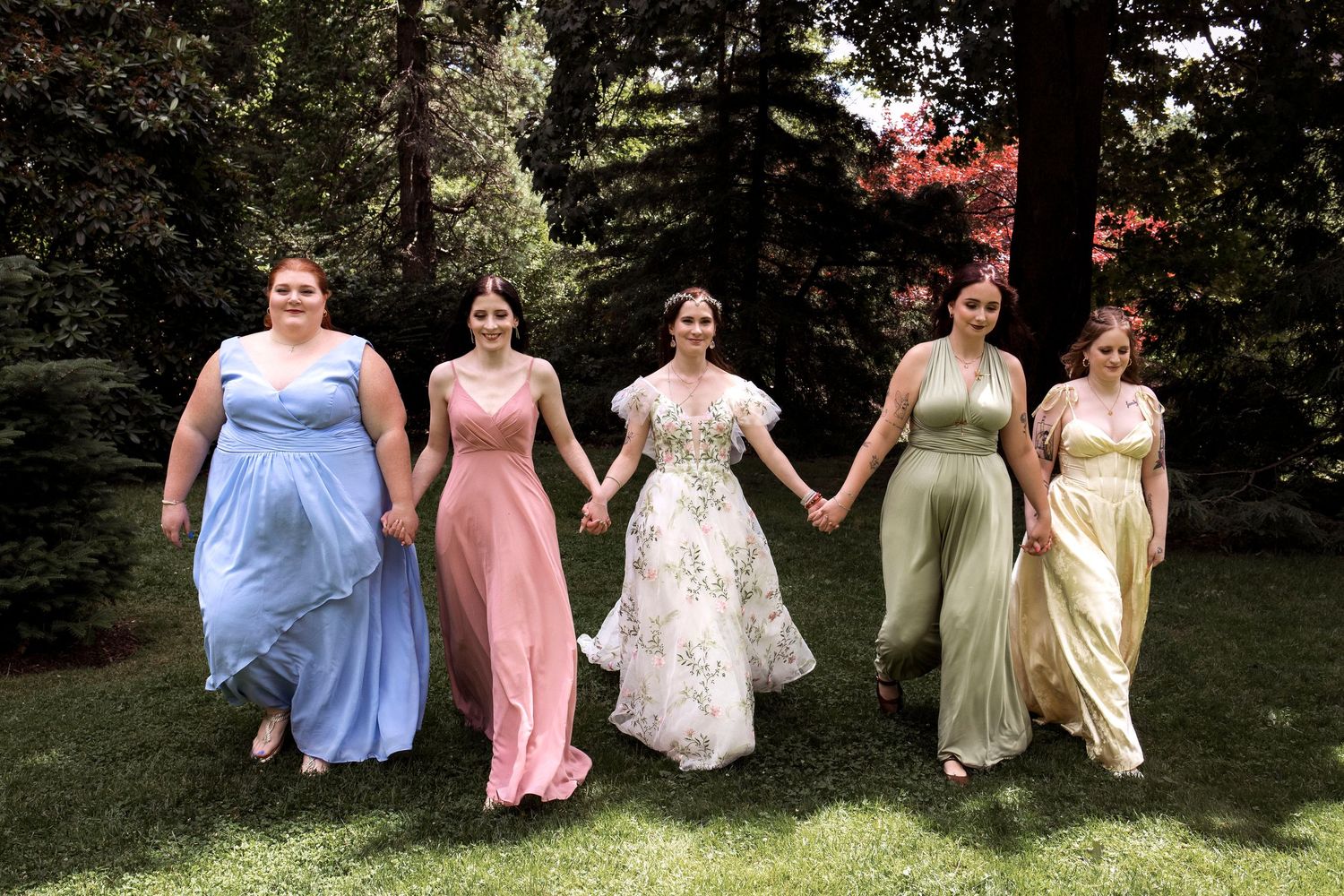 Bride in floral gown holds hands with four bridesmaids in colorful pastel dresses, walking on green lawn outdoors.