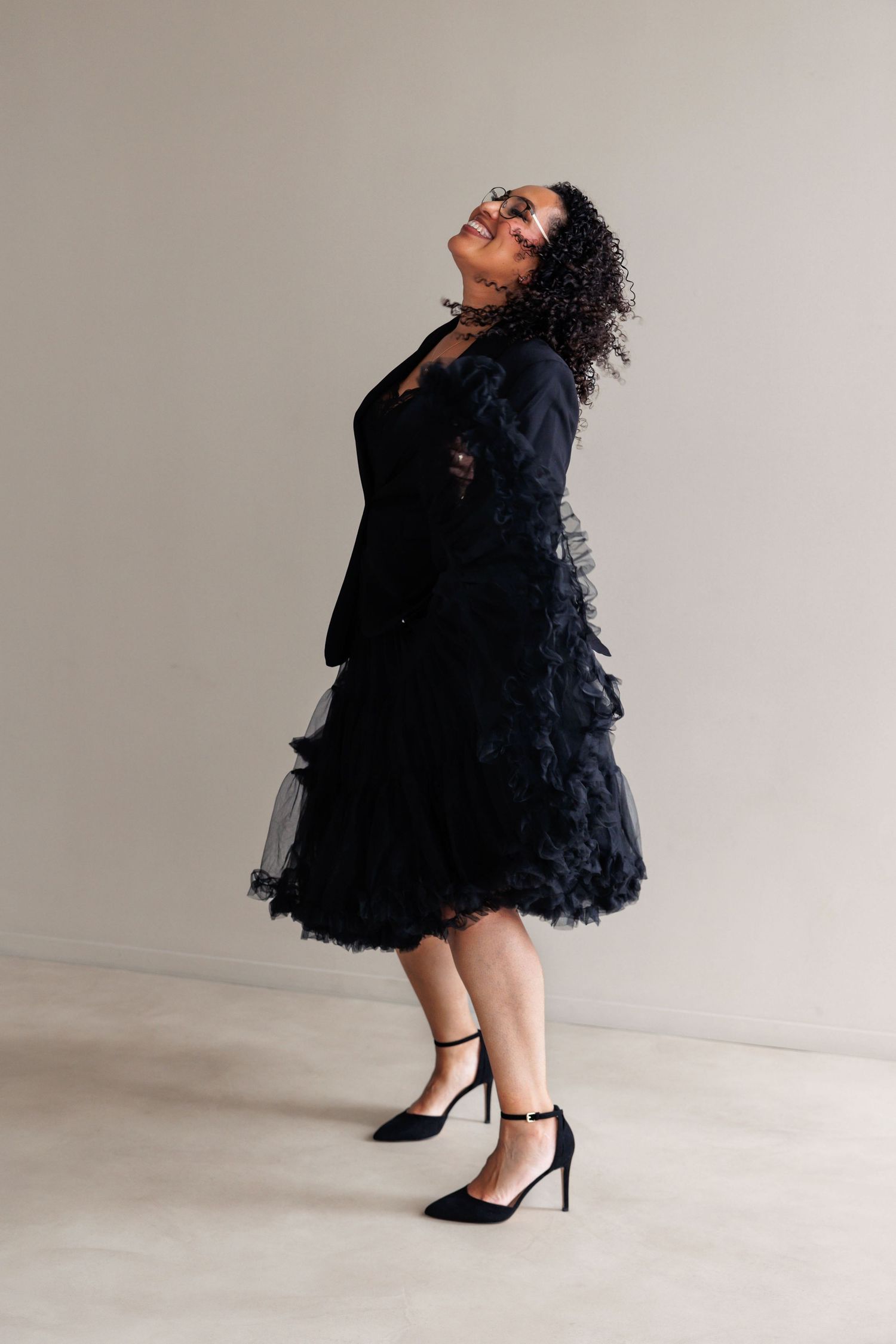 Woman in black ruffled tulle skirt, blazer, and heels posing confidently against a neutral background.