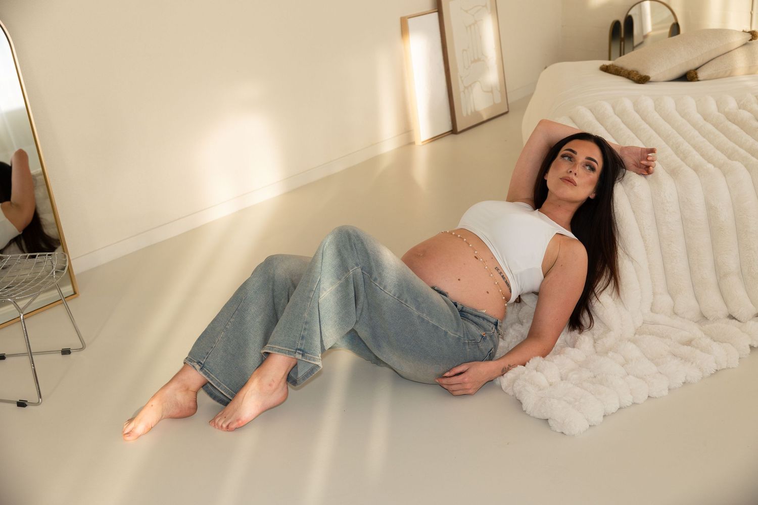 Pregnant woman in white crop top and jeans relaxing on floor, showcasing baby bump in bright minimalist room.