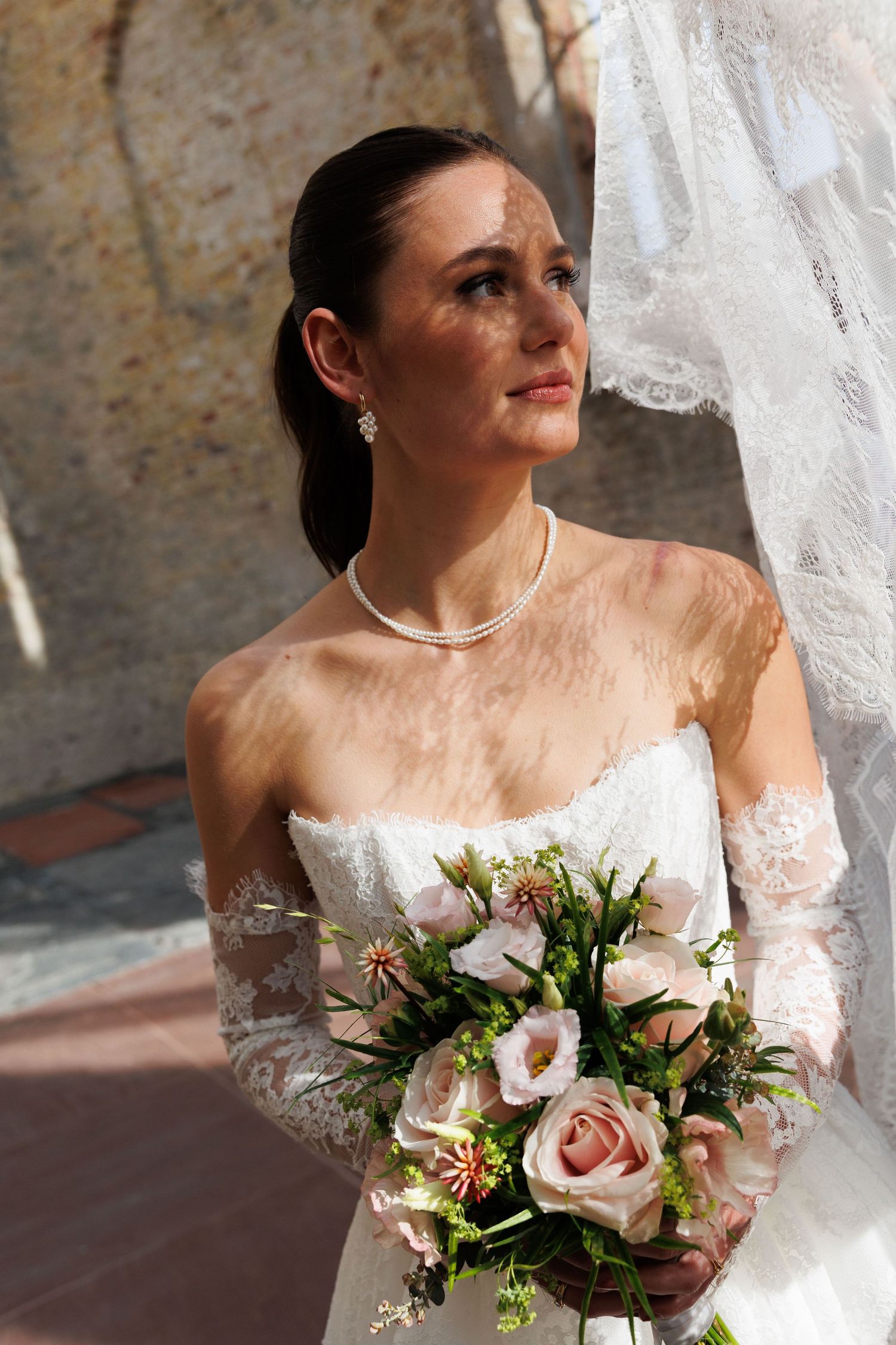 Elegant bride in white lace gown holding pink rose bouquet, wearing pearl necklace and veil outdoors.