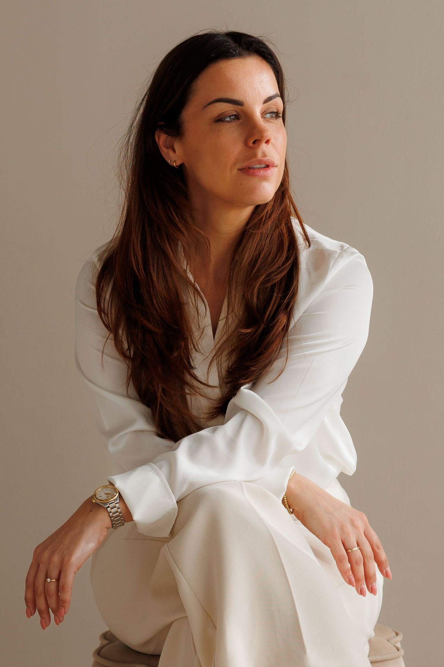 Woman in white blouse and trousers sitting gracefully, gazing away thoughtfully, wearing a gold watch.