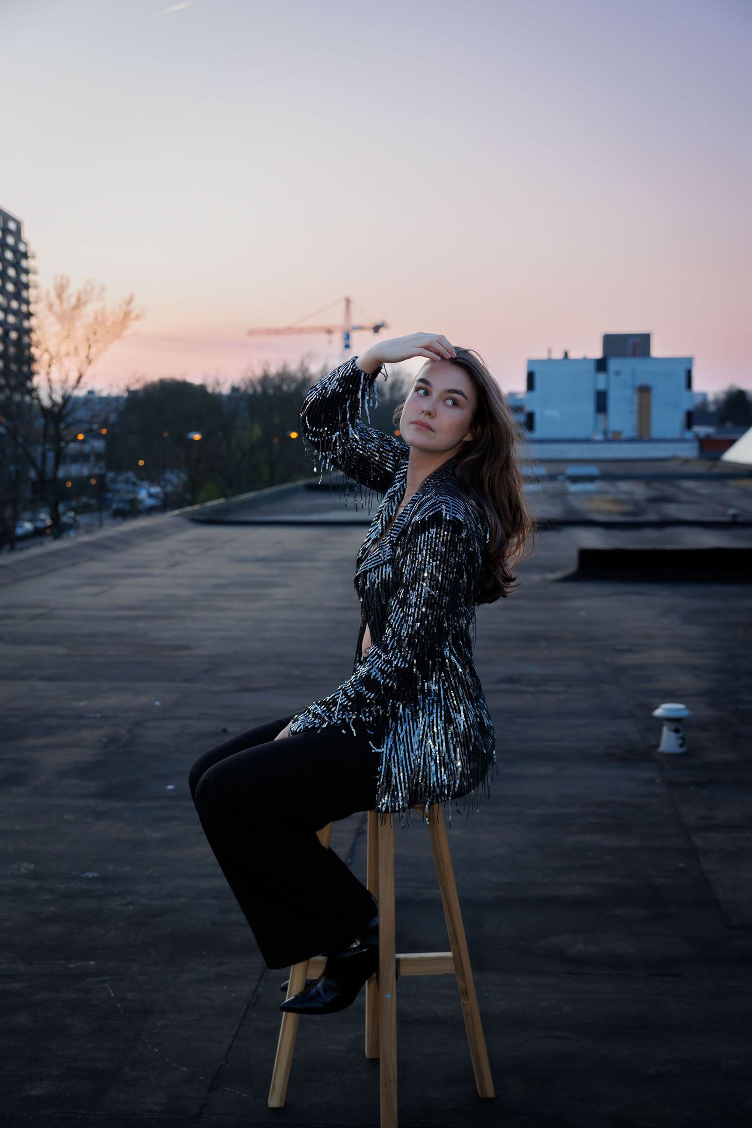 Young woman in sequin jacket poses on stool atop urban rooftop during pink sunset, with city buildings behind her.