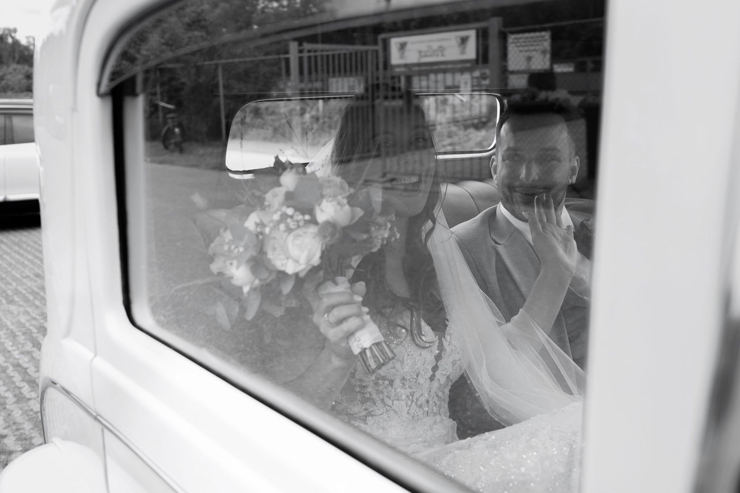 Black and white photo of a bride and groom sitting in a vintage white car, bride holding a bouquet.