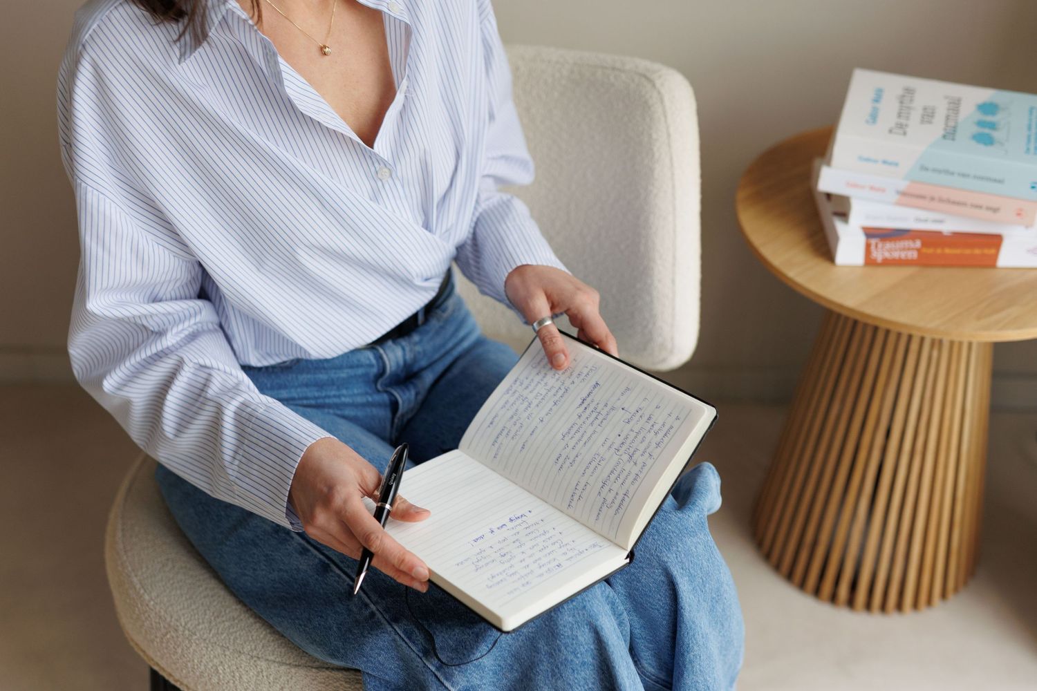 Woman in white striped shirt and jeans sitting on chair reading a notebook with pen in hand near wooden side table.