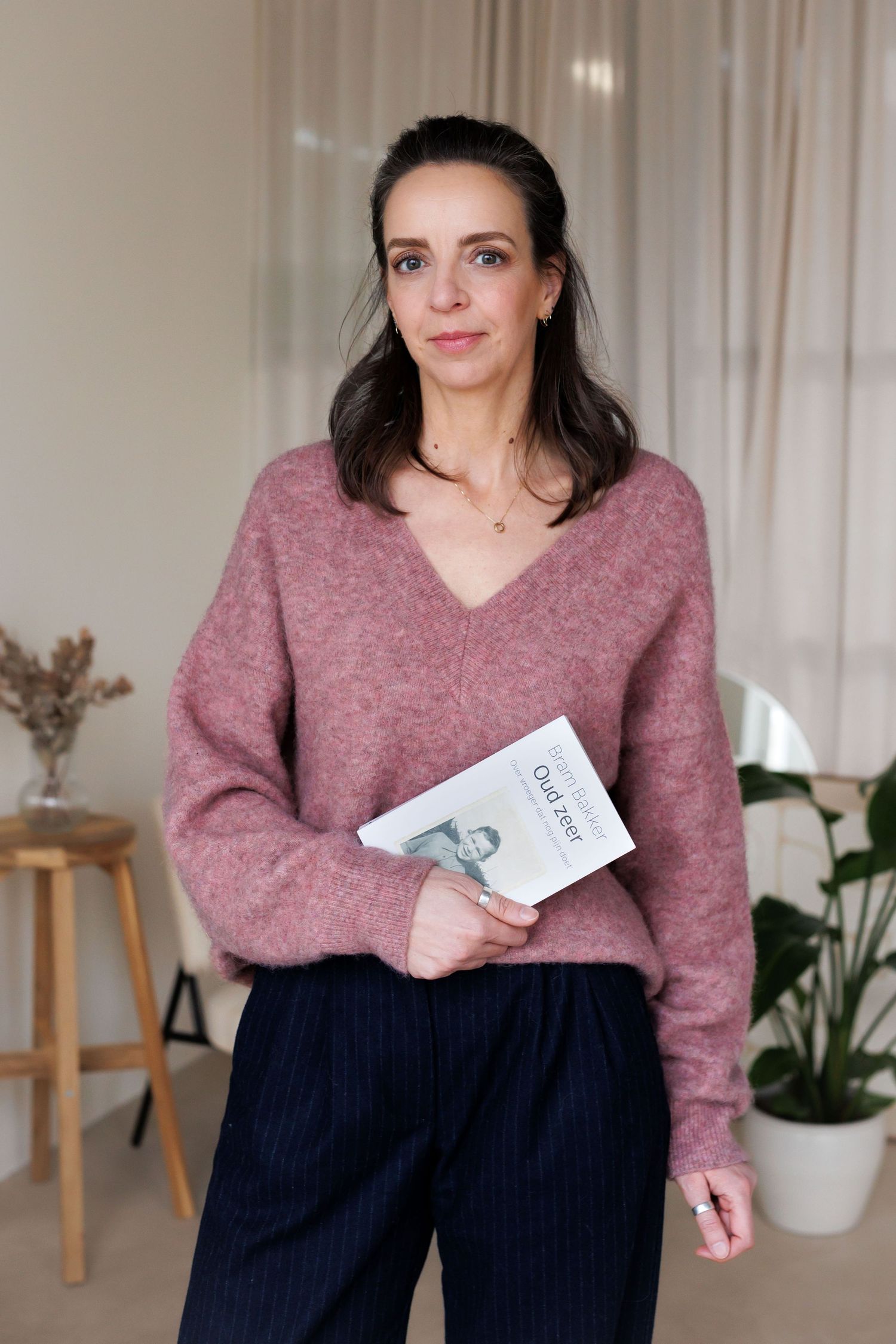 Woman in pink fuzzy V-neck sweater holding a book, standing in a bright modern room with plants.