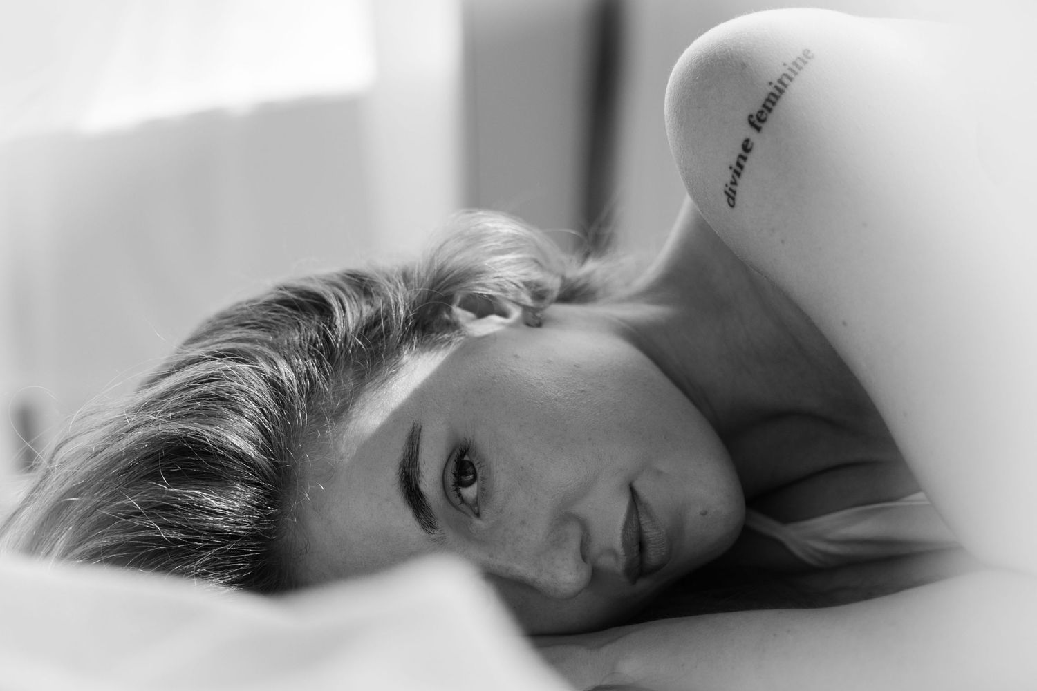 Black and white portrait photograph capturing someone resting on white bedsheets with natural lighting.