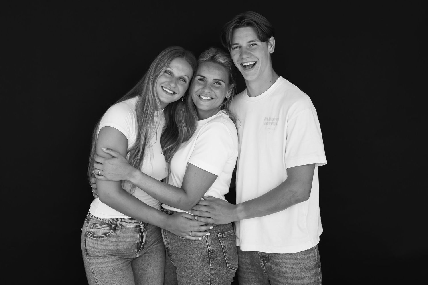 Black and white photo of three smiling young adults in white tees and jeans posing together against a dark background.