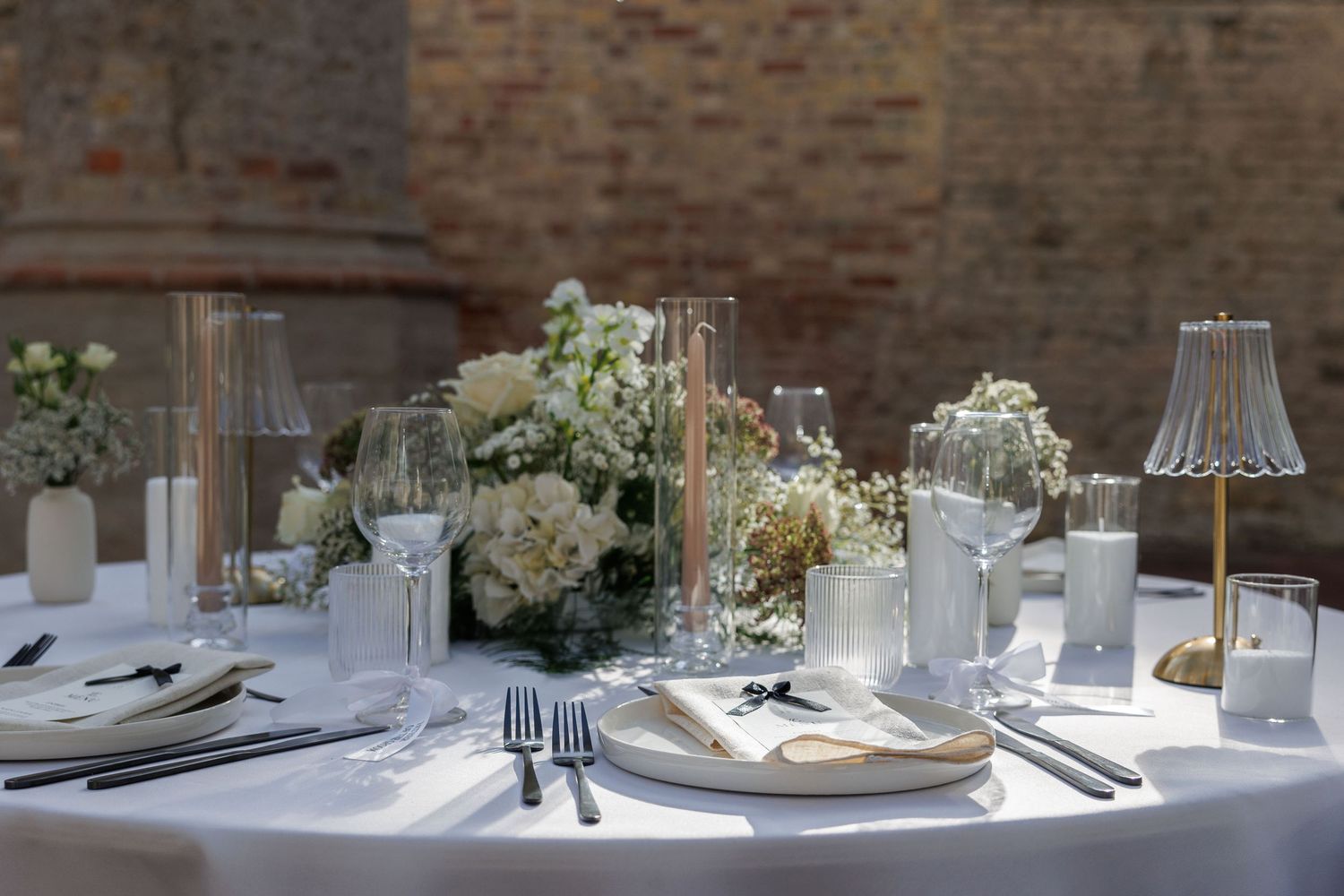 Elegant wedding reception table with white floral centerpiece, candles, crystal glassware, and white linens in a rustic venue.