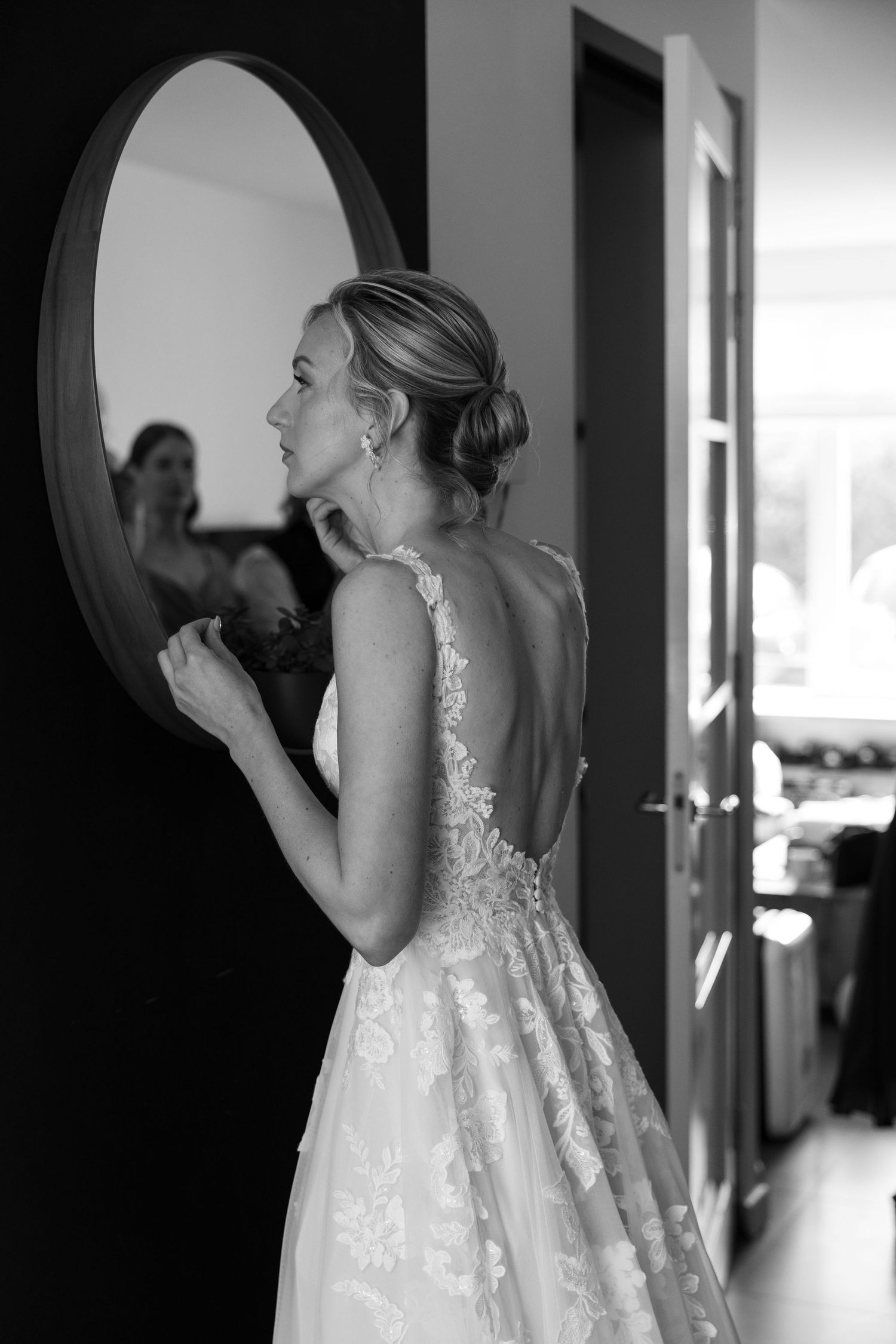 Black and white photo of a bride in a backless lace wedding gown gazing into a round mirror while getting ready.